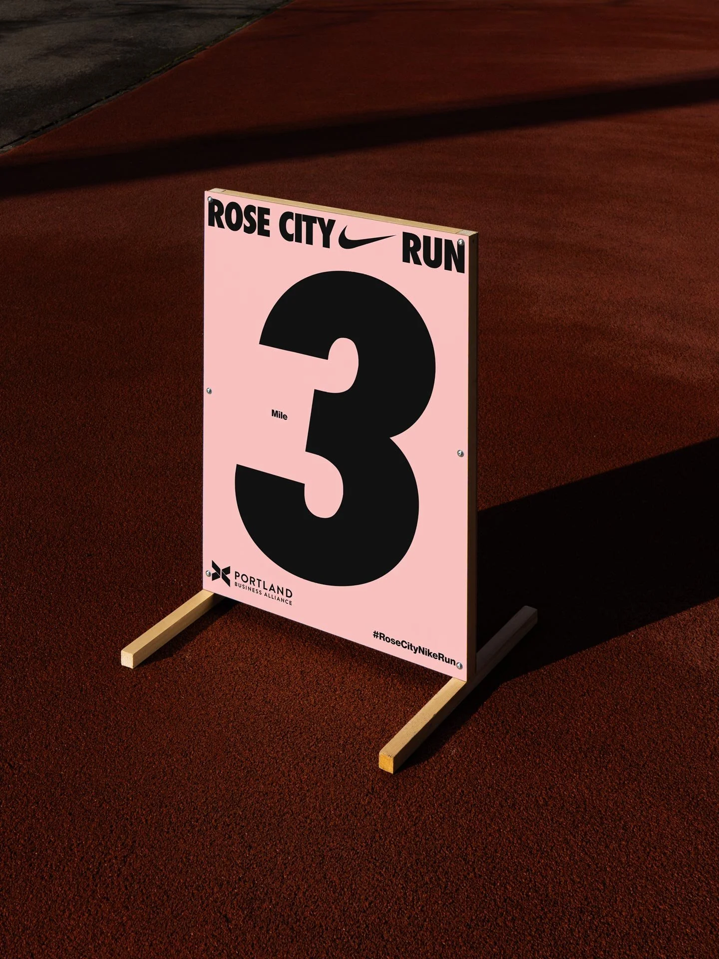 Pink race sign for Rose City Nike Run, indicating 3 miles, placed on a running track with sunlight casting a shadow.