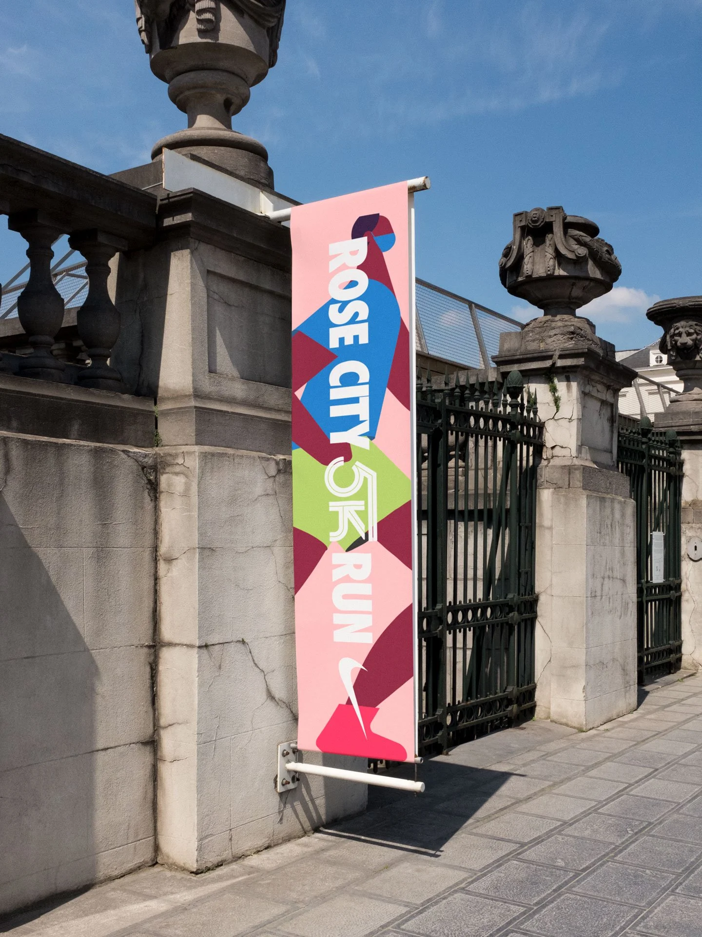 Colorful vertical banner with abstract geometric design and text reading 'Rose City 5K Run', mounted on a stone wall near a metal gate under a blue sky.
