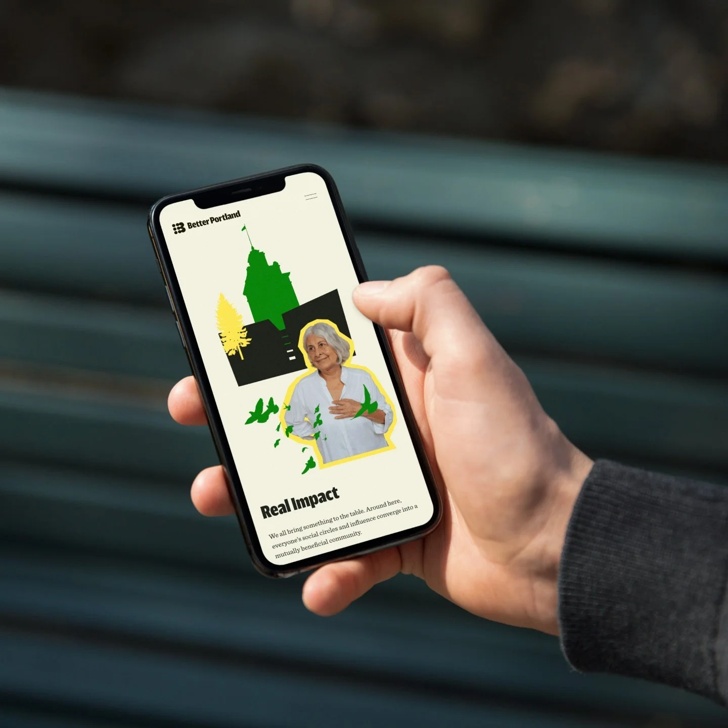 Close-up of a hand holding a smartphone showing a webpage titled 'Better Portland' with images of a city skyline, a woman, and green birds, and the heading 'Real Impact'.
