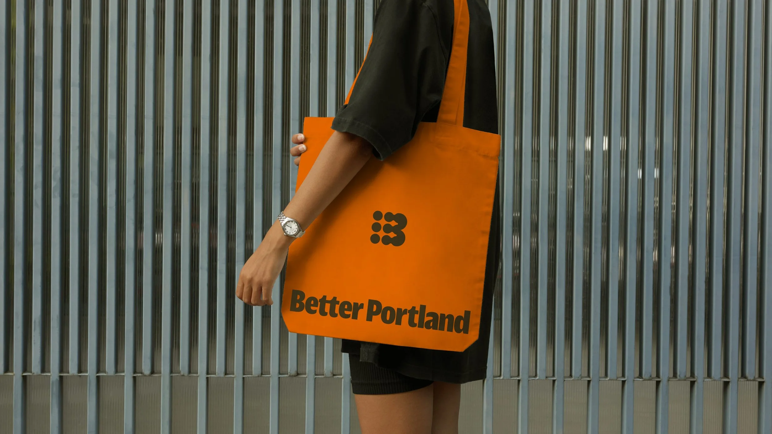 Person in black clothing carrying an orange tote bag with the logo and text "Better Portland" written on it, standing in front of a metallic, vertical-paneled wall.