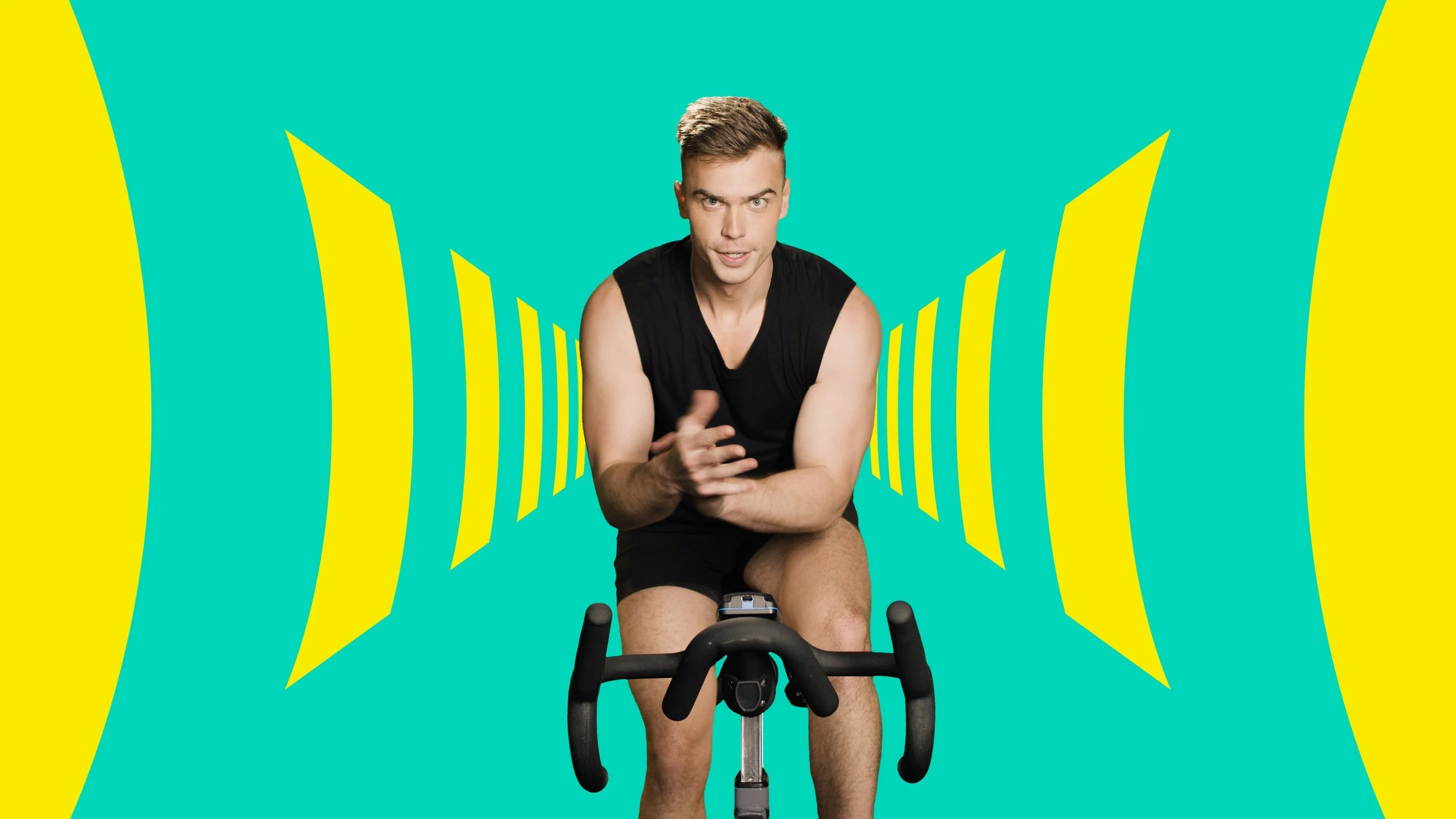 A young man in black athletic wear riding a stationary exercise bike in front of a colorful, abstract background with yellow and teal shapes.