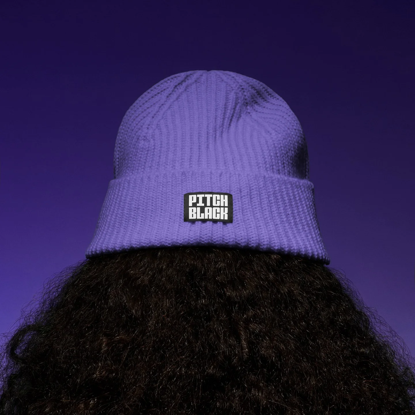 A purple knit beanie with a black and white 'Pitch Black' patch on the front, worn over long dark curly hair against a purple gradient background.