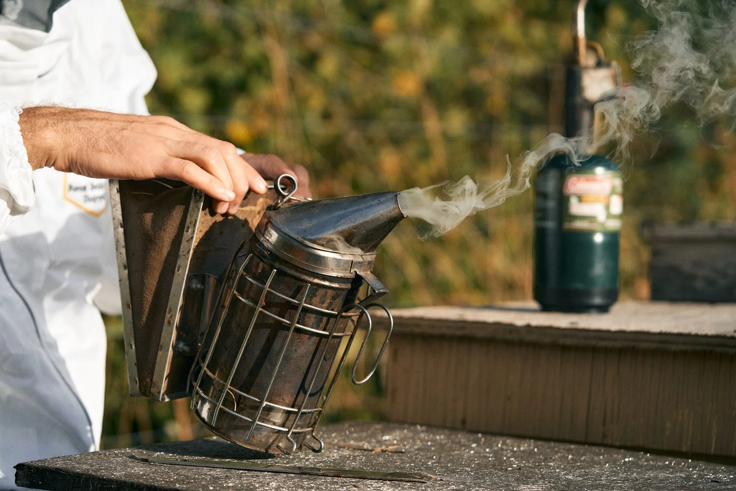 A person wearing a white protective suit is handling a homemade smoke generator or device that emits white smoke or vapor outdoors. The device is a metal canister with a cone-shaped top and a wire cage around it, placed on a wooden surface. A gas canister is visible in the background.