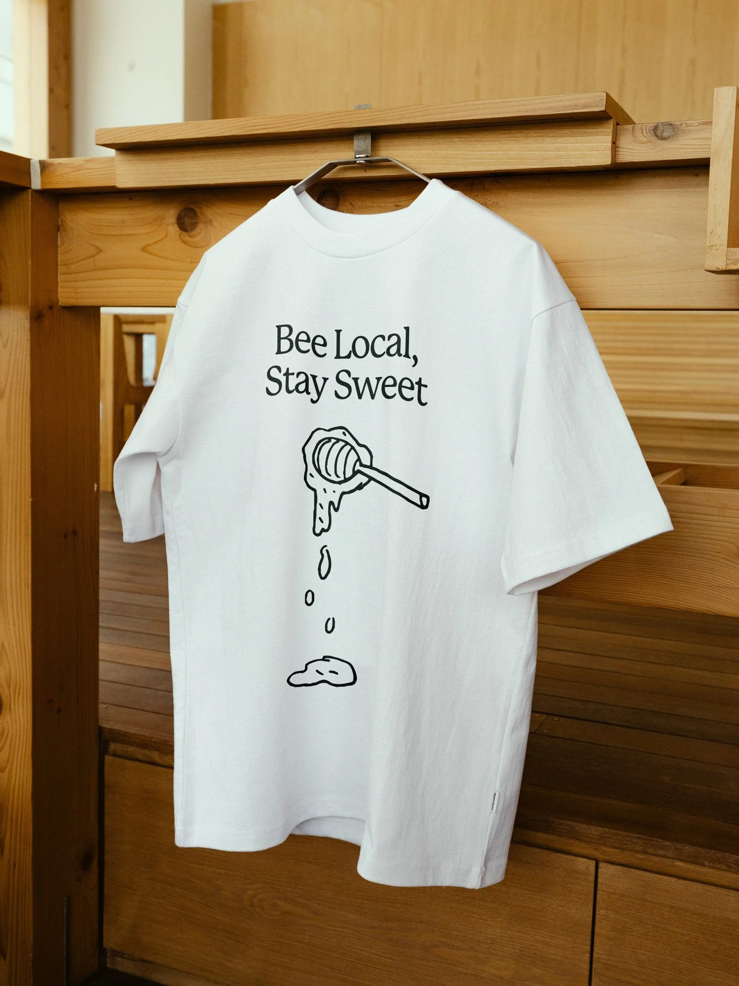 White T-shirt hanging on a hanger with the text 'Bee Local, Stay Sweet' and a graphic of a honey dipper dripping honey.