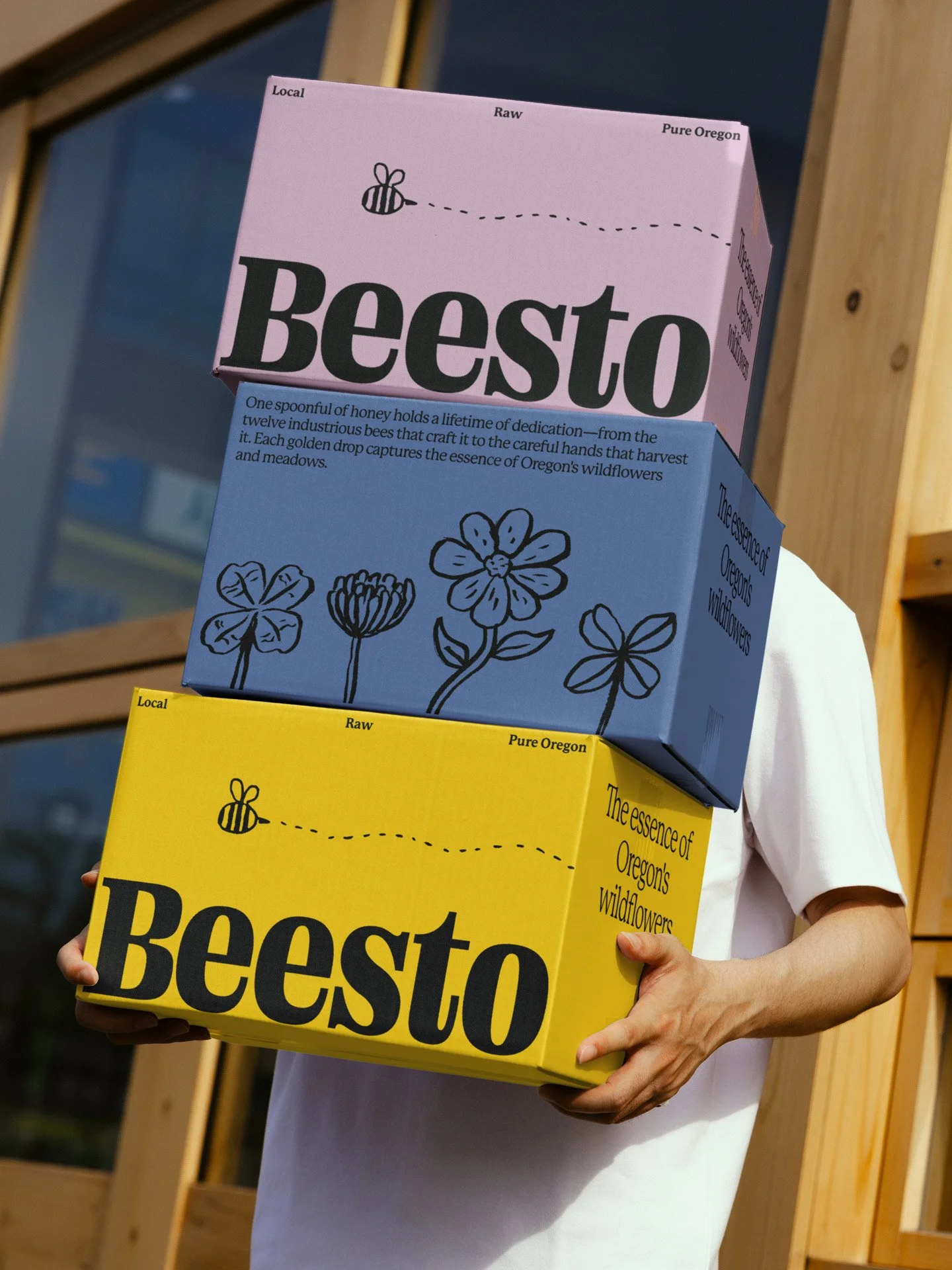 Person holding two colorful boxes labeled Beesto, one pink and one yellow, with illustrations of flowers and a bee icon, against a background of wooden structures.