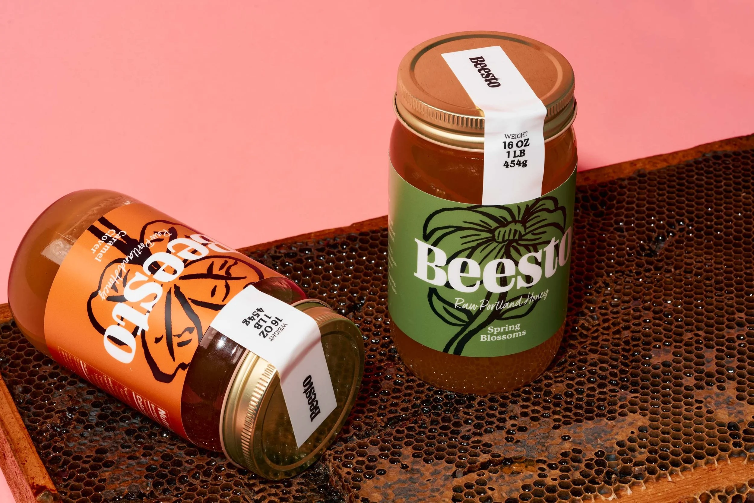 Three jars of beeswax lip balm on a honeycomb. One jar is upright with a green label that says 'Beesto' and 'Spring Blossoms.' Two jars are fallen on their sides, one with an orange label and the other with a white label.