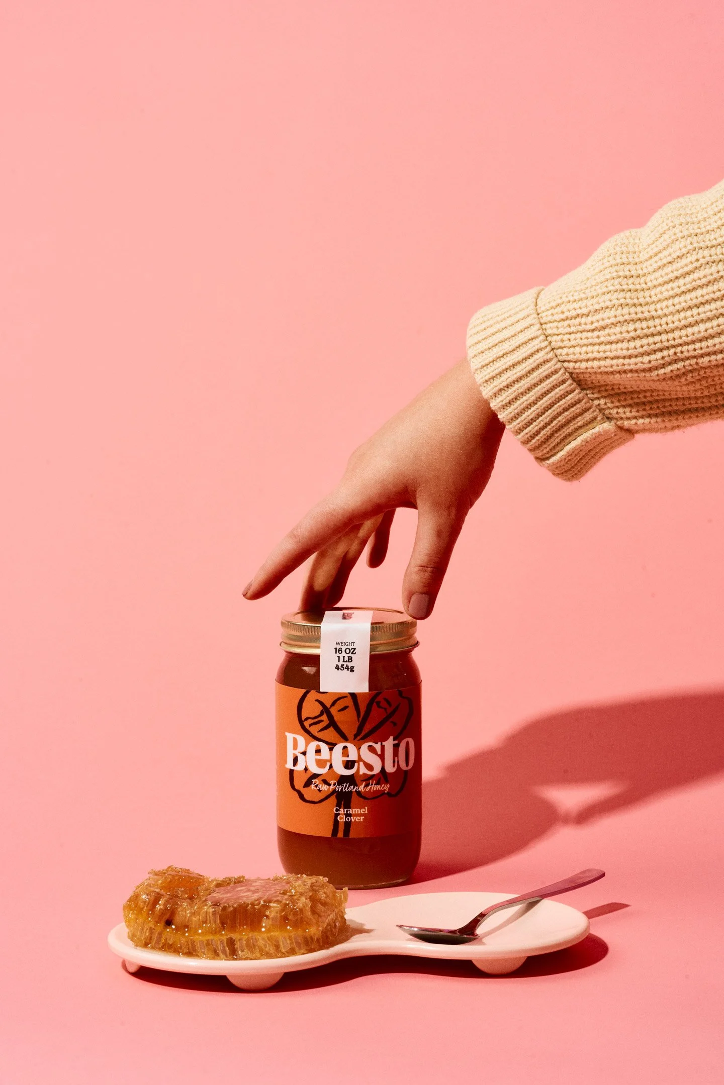 A hand wearing a beige knit sleeve reaching to open a jar of Beesto caramel clover honey against a pink background, with a white plate holding caramel honeycomb, a spoon, and a plastic knife.