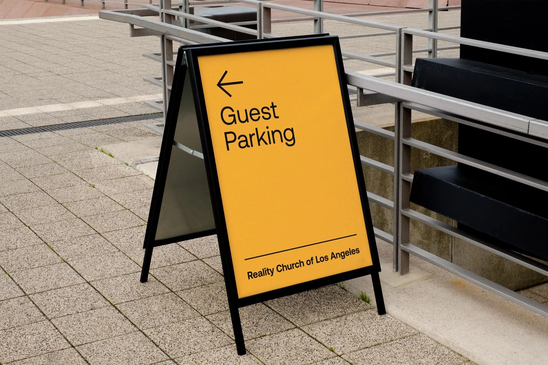 A yellow sidewalk sign indicating guest parking to the left, with the text 'Reality Church of Los Angeles' at the bottom, placed on a sidewalk with metal railing and bench nearby.