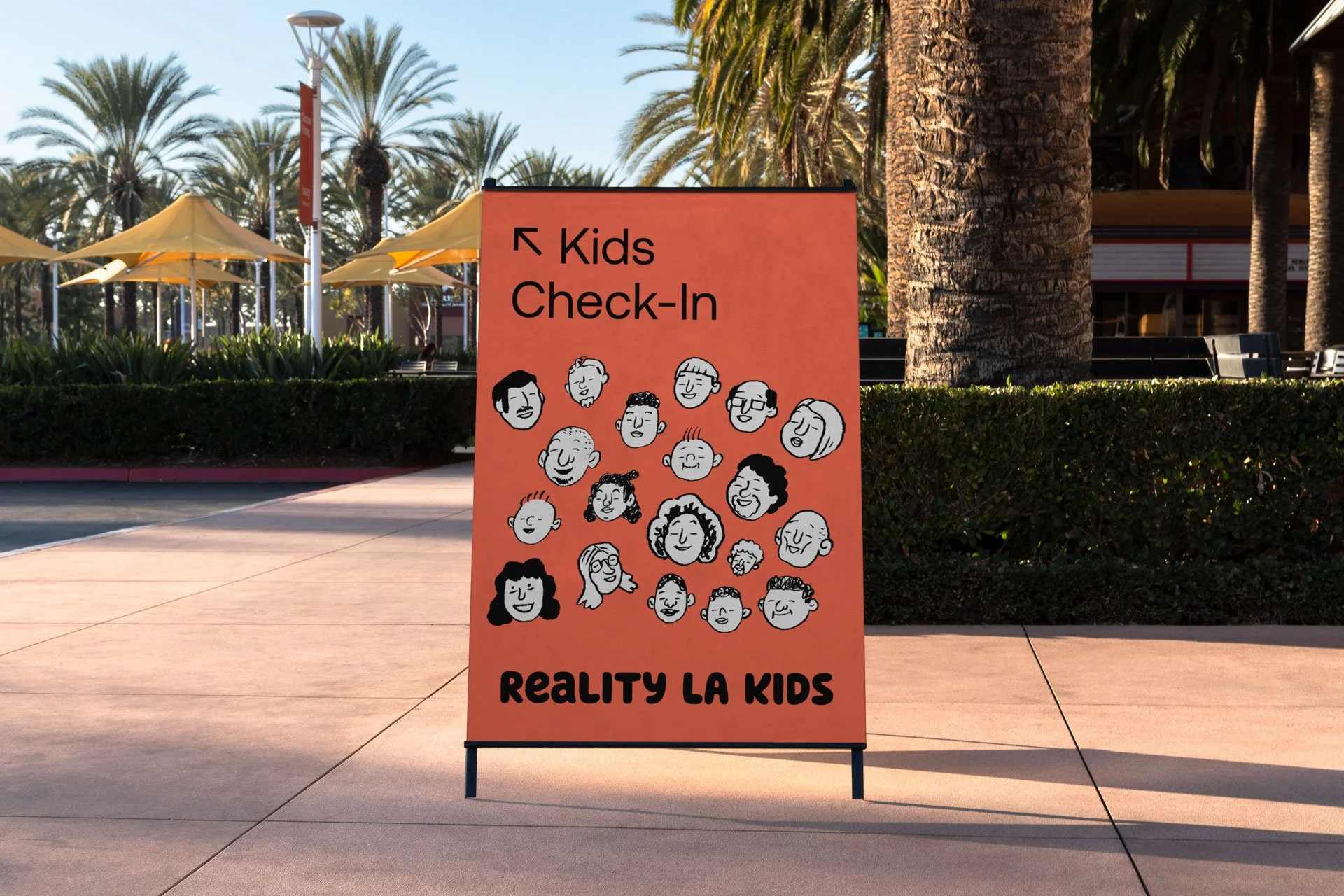 Bright pink sign with cartoon faces and text for Kids Check-In at Reality LA Kids, positioned outdoors with palm trees and yellow umbrellas in the background.