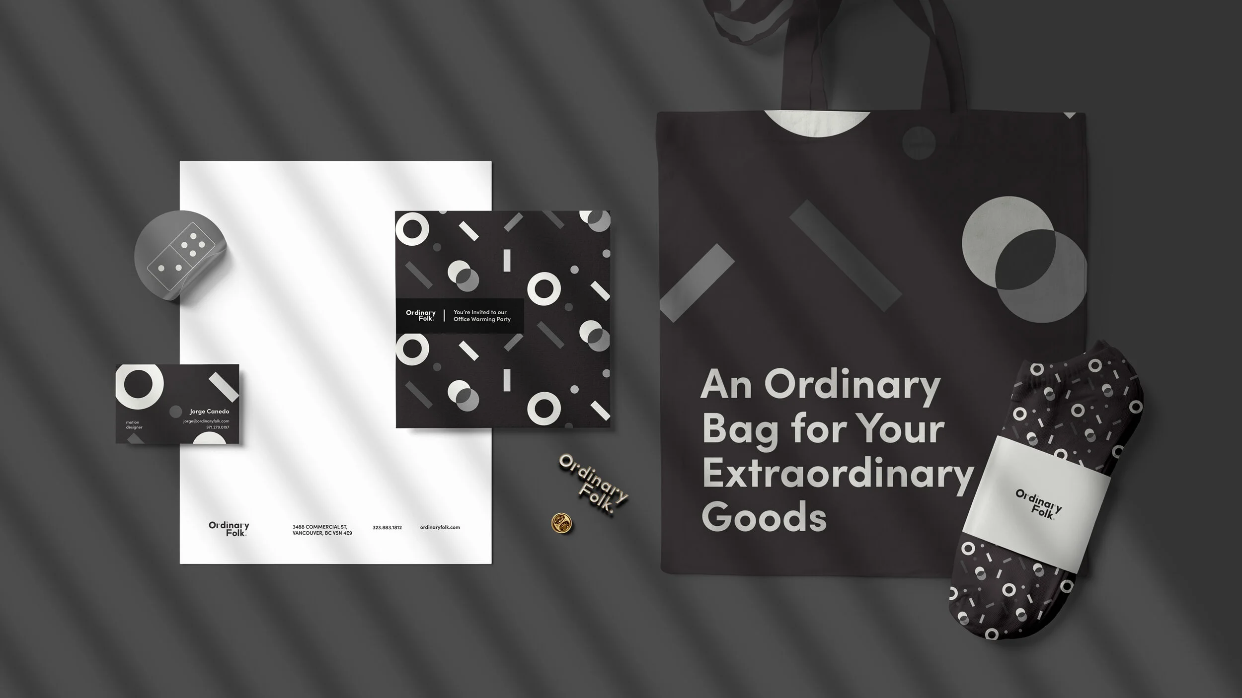 Black tote bag with white text that reads 'An Ordinary Bag for Your Extraordinary Goods,' alongside branded stationery items, business card, and wrapped package with circular and geometric patterns in black, white, and gray.