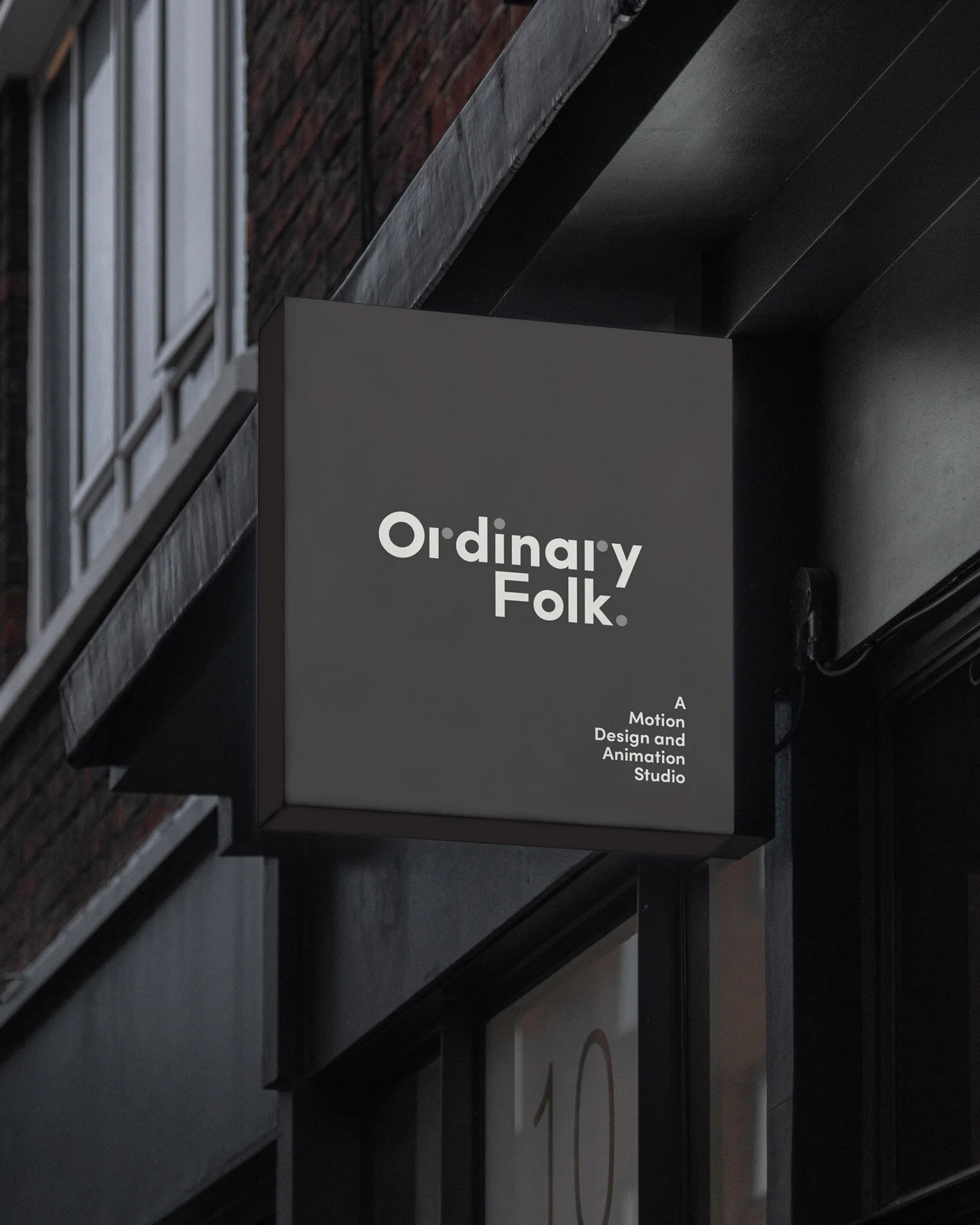 A black and gray sign with the text "Ordinary Folk." and smaller text below that reads "A Motion Design and Animation Studio." The sign is attached to the wall outside a building with brick and metal siding, under a dark awning.