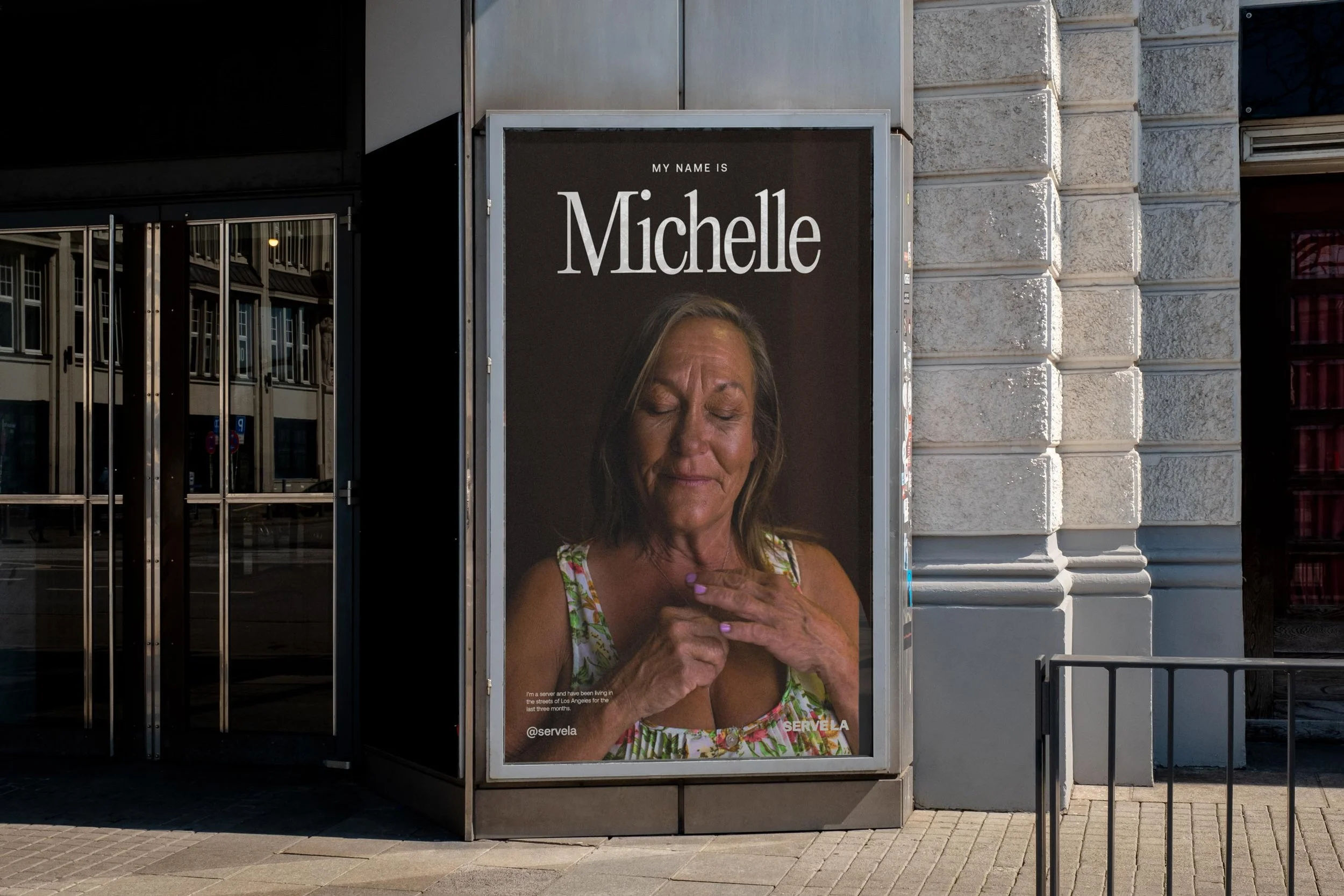 A woman smiling and looking down at her chest with her eyes closed, wearing a floral dress, stands in front of a large advertisement display that says "My name is Michelle" and includes her name and part of her story. The display is on a city sidewalk, next to a stone building with glass doors.