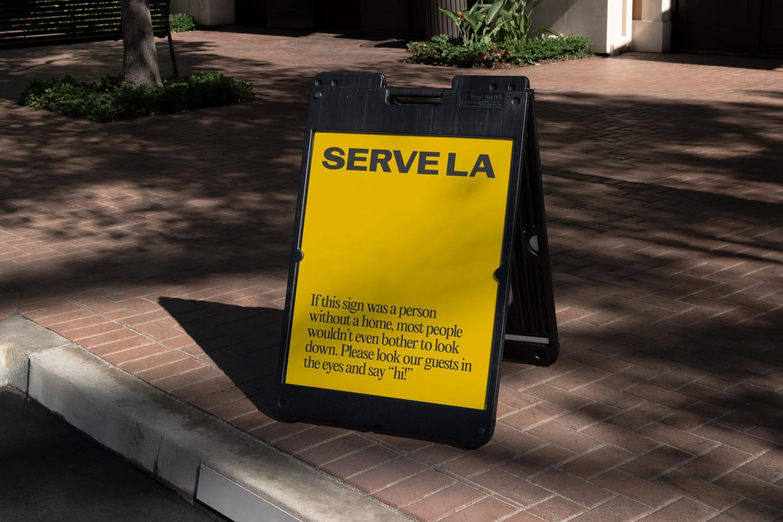 A yellow and black sidewalk sign with the word 'SERVE LA' on top, placed on a brick sidewalk near some plants and a tree. The sign contains a message inviting guests to greet others with eye contact and a smile.