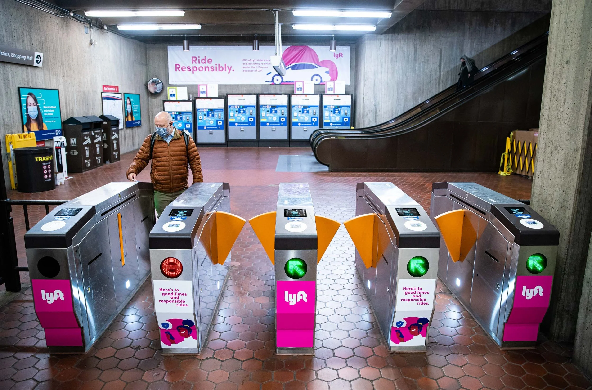 A subway or train station entrance with turnstiles for ticketing. There are orange barriers with digital card readers on top. To the left, there are trash and recycling bins, and a man wearing a brown jacket and face mask is passing through the turnstile. In the background, there are electronic displays and advertisements, including a large pink sign with the Lyft logo and slogan, and escalators on the right side with a woman riding them.