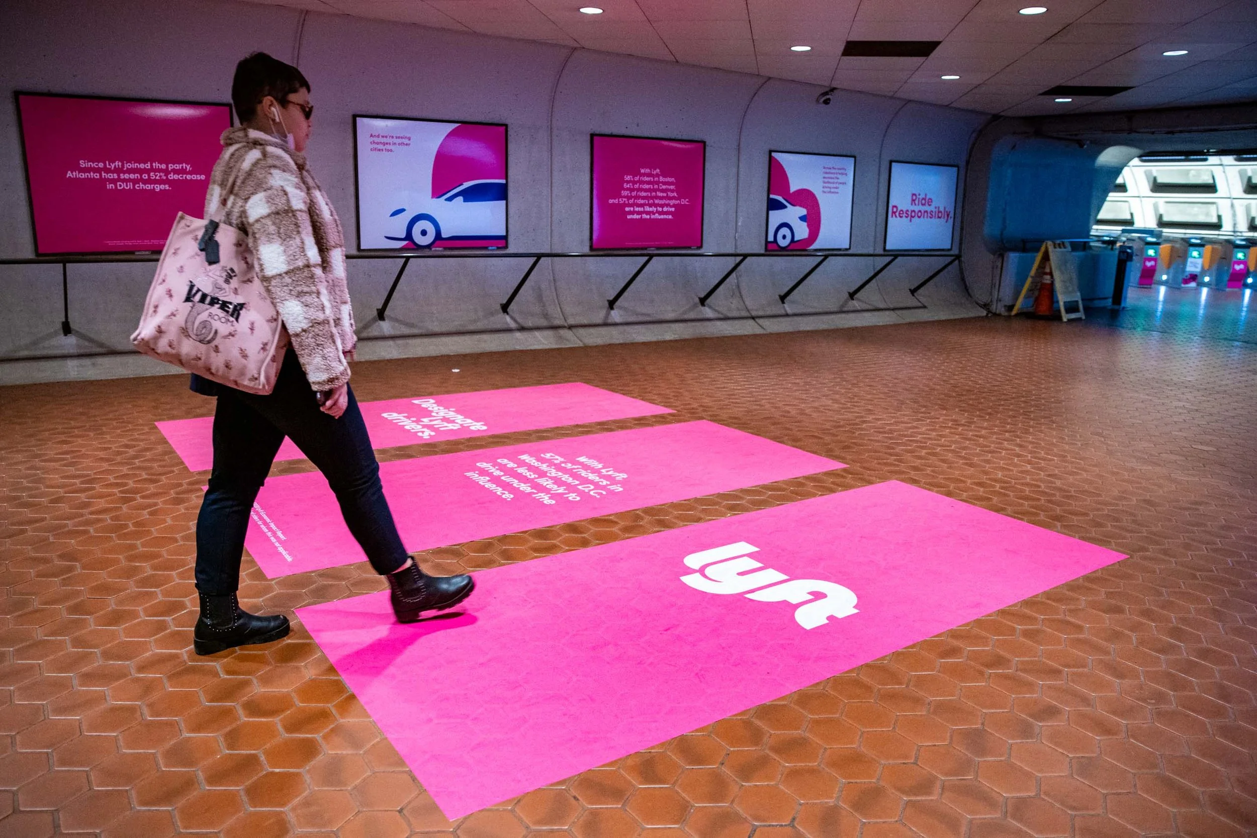 A woman walking in an underground station with pink and white digital advertisements on the wall and pink floor markings, wearing glasses, a checkered coat, and carrying a pink bag that has the word 'VIRFLY' on it.