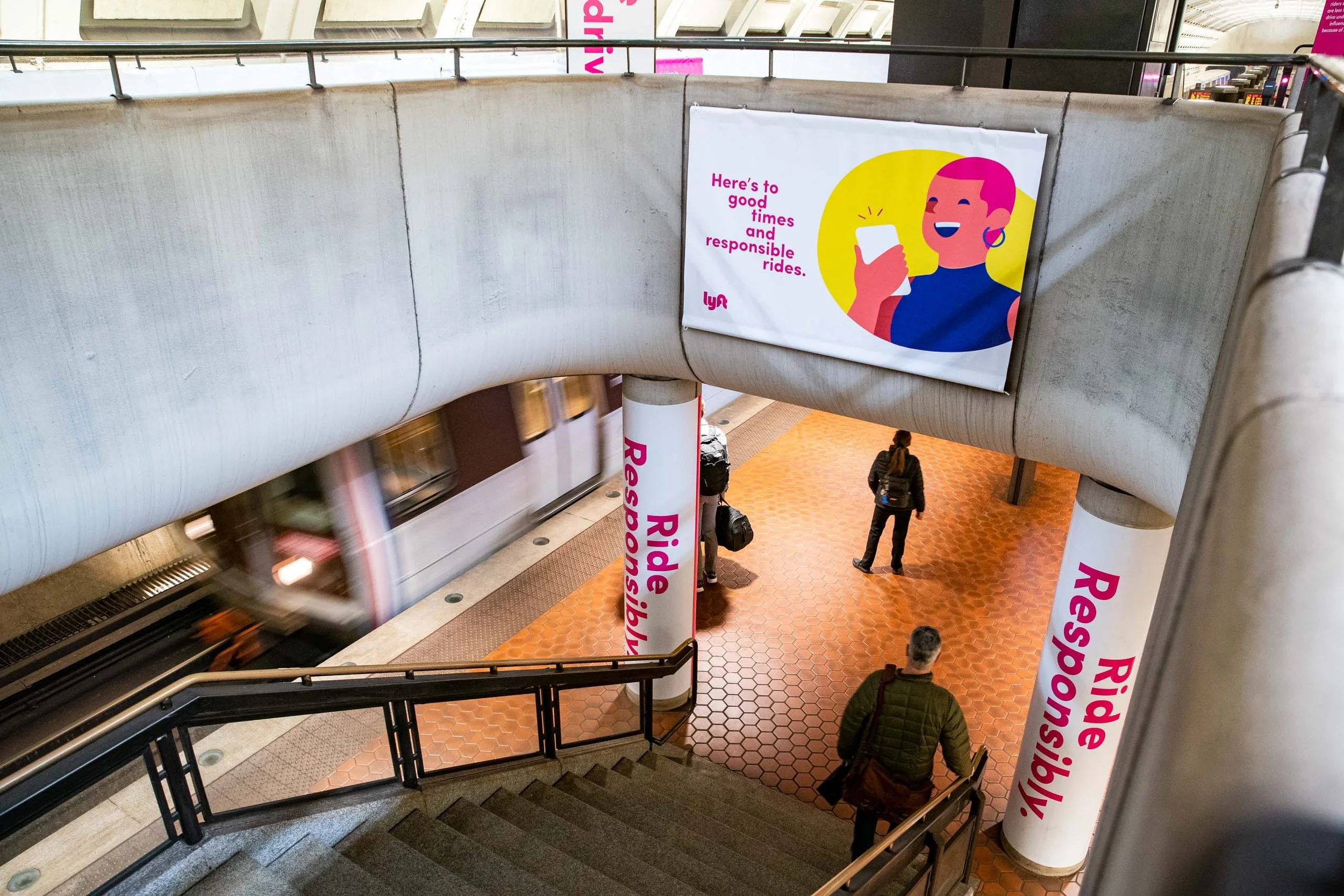 People walking in a subway station with pink and white Lyft banners, escalators, and tiled flooring.