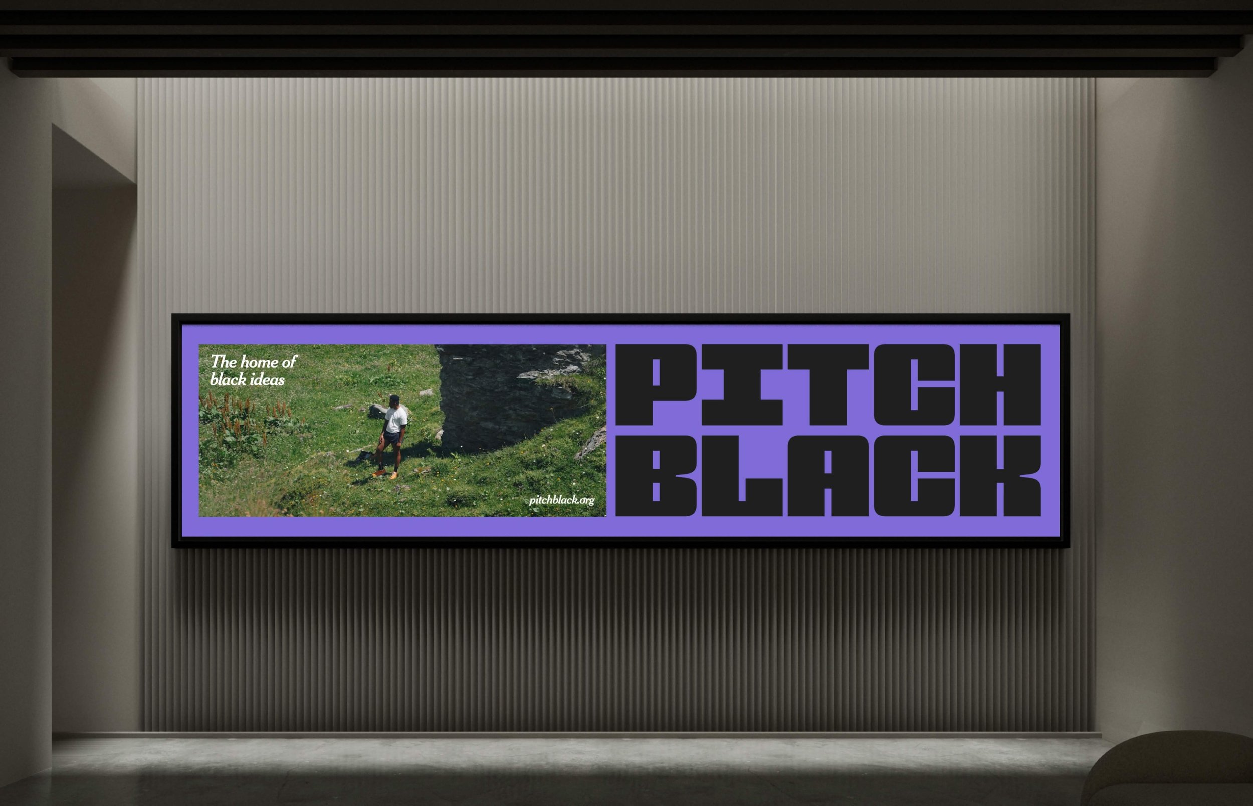 A digital billboard with a lavender border displays a photo of a person standing on grass near a large rock, with a text overlay that says "The home of black ideas" and a website URL pitchblack.org. The billboard is mounted on a wall with vertical ribbed paneling, and the room has a modern, minimalist style.