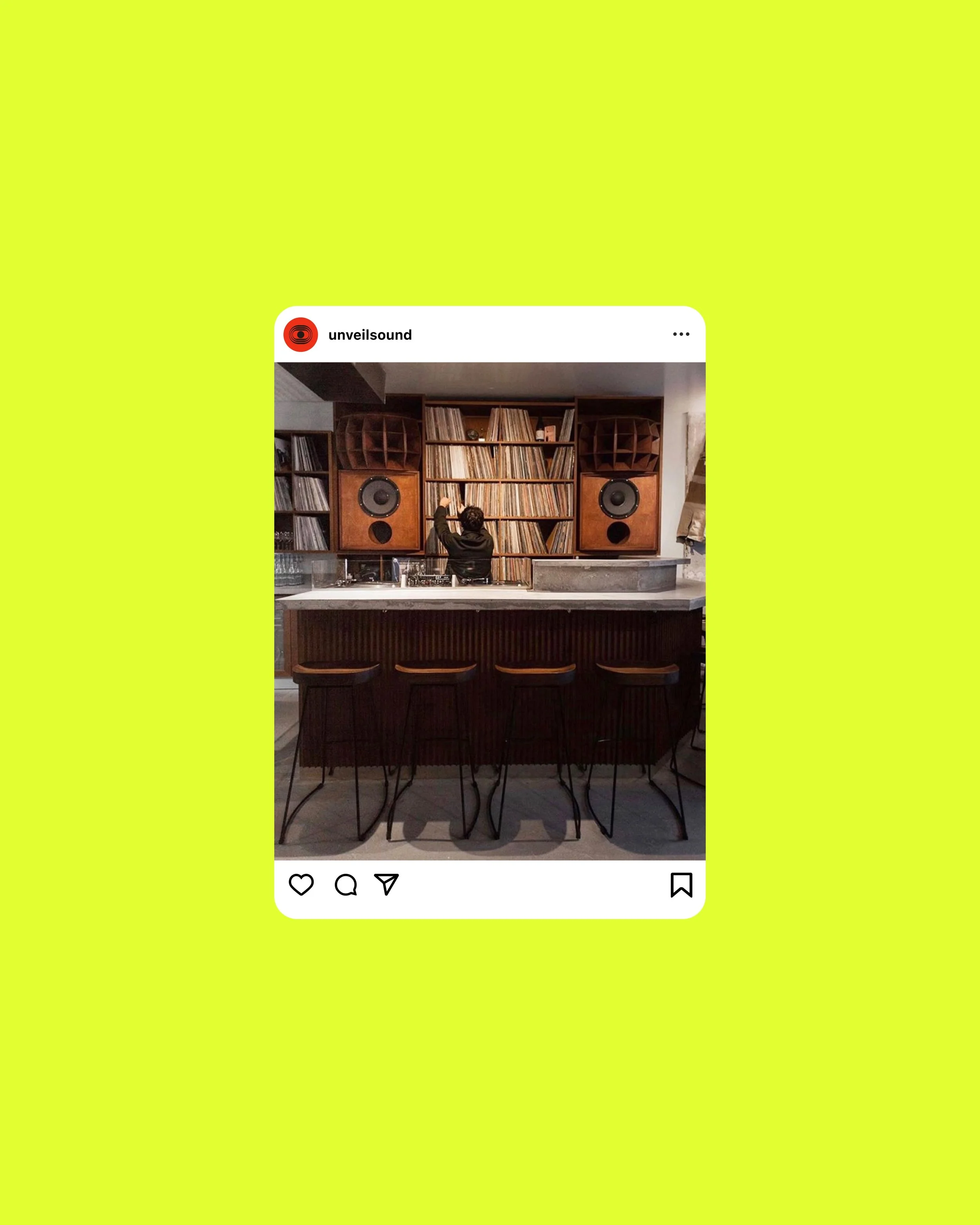 Interior of a restaurant or bar with a wooden counter and high chairs, a large wooden bookshelf filled with vinyl records, and two vintage speakers on either side of the bookshelf.