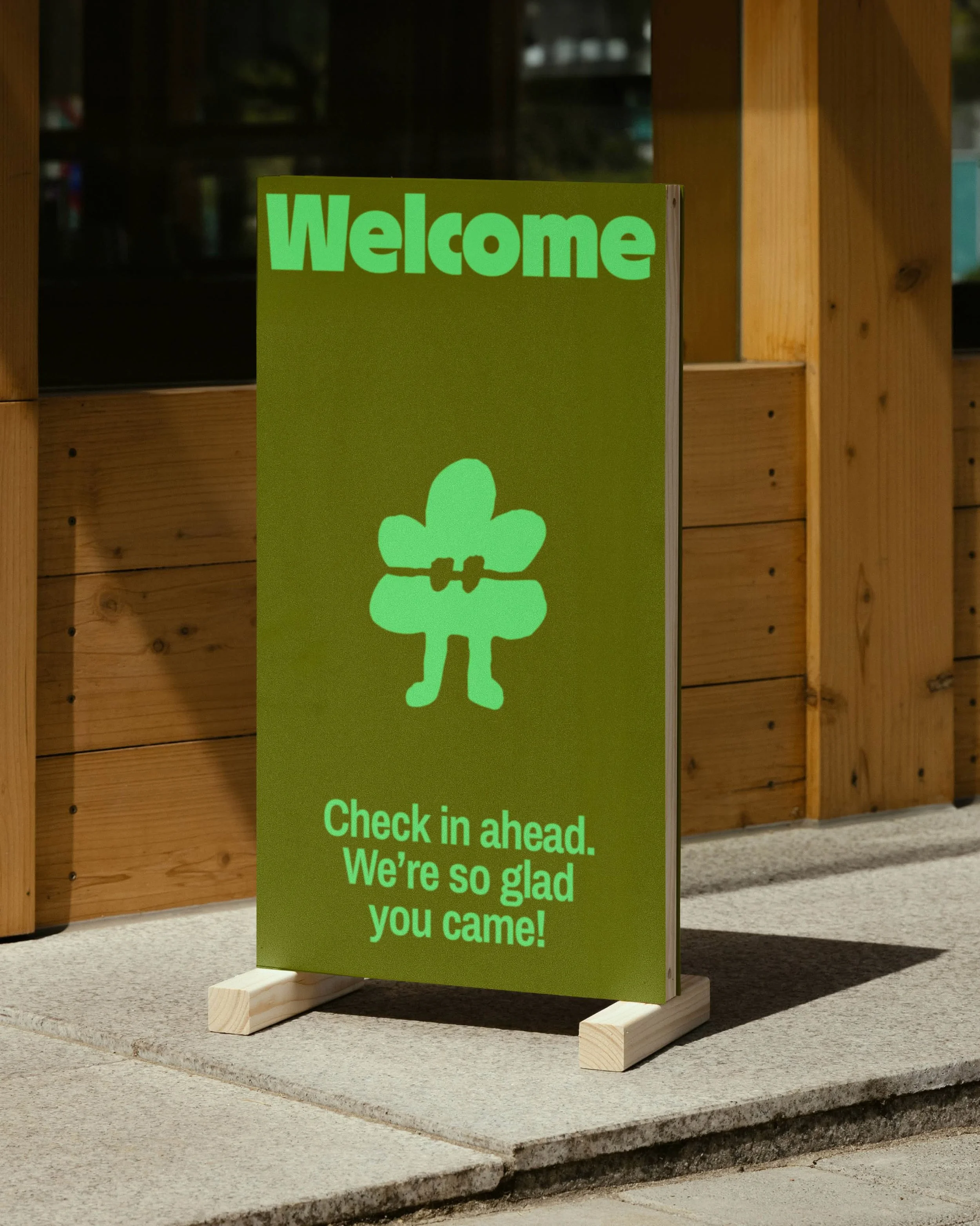 Green welcome sign with a clover symbol and text that says 'Check in ahead. We're so glad you came!' placed on a wooden stand outside a building.