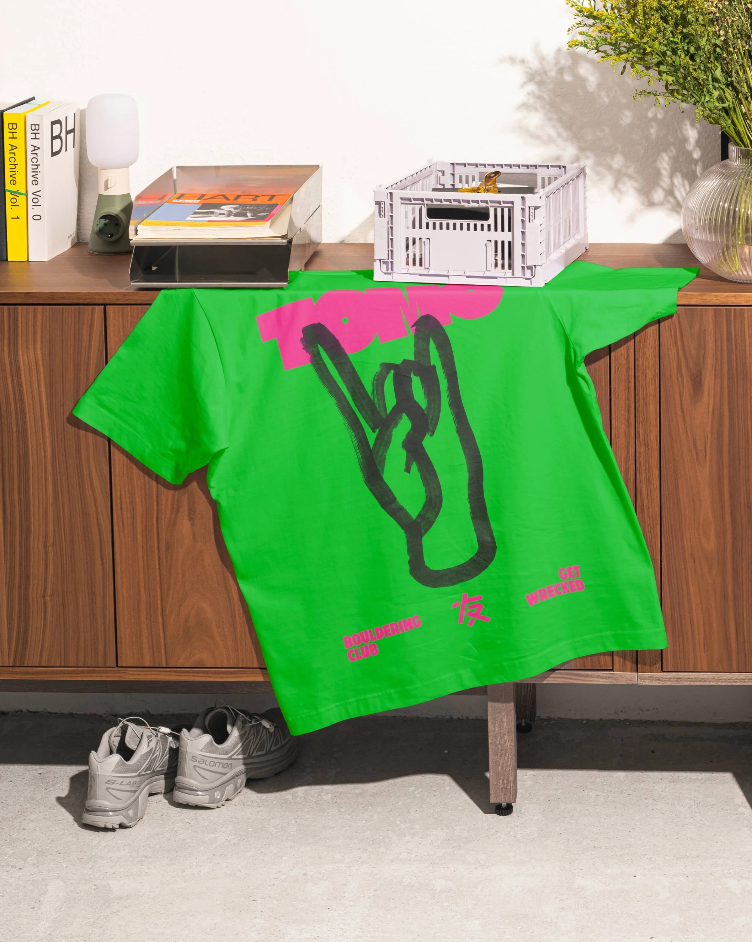 A wooden table with a bright green banner hanging from it that reads 'Bouldering Club' in pink letters with some Chinese characters underneath. On the table are a pile of magazines, a gray and white lamp, and a white plastic crate with a bottle opener on top. On the ground below the table are a pair of white Skechers athletic shoes. The background is a plain white wall with part of a glass vase with green plants on the right side.
