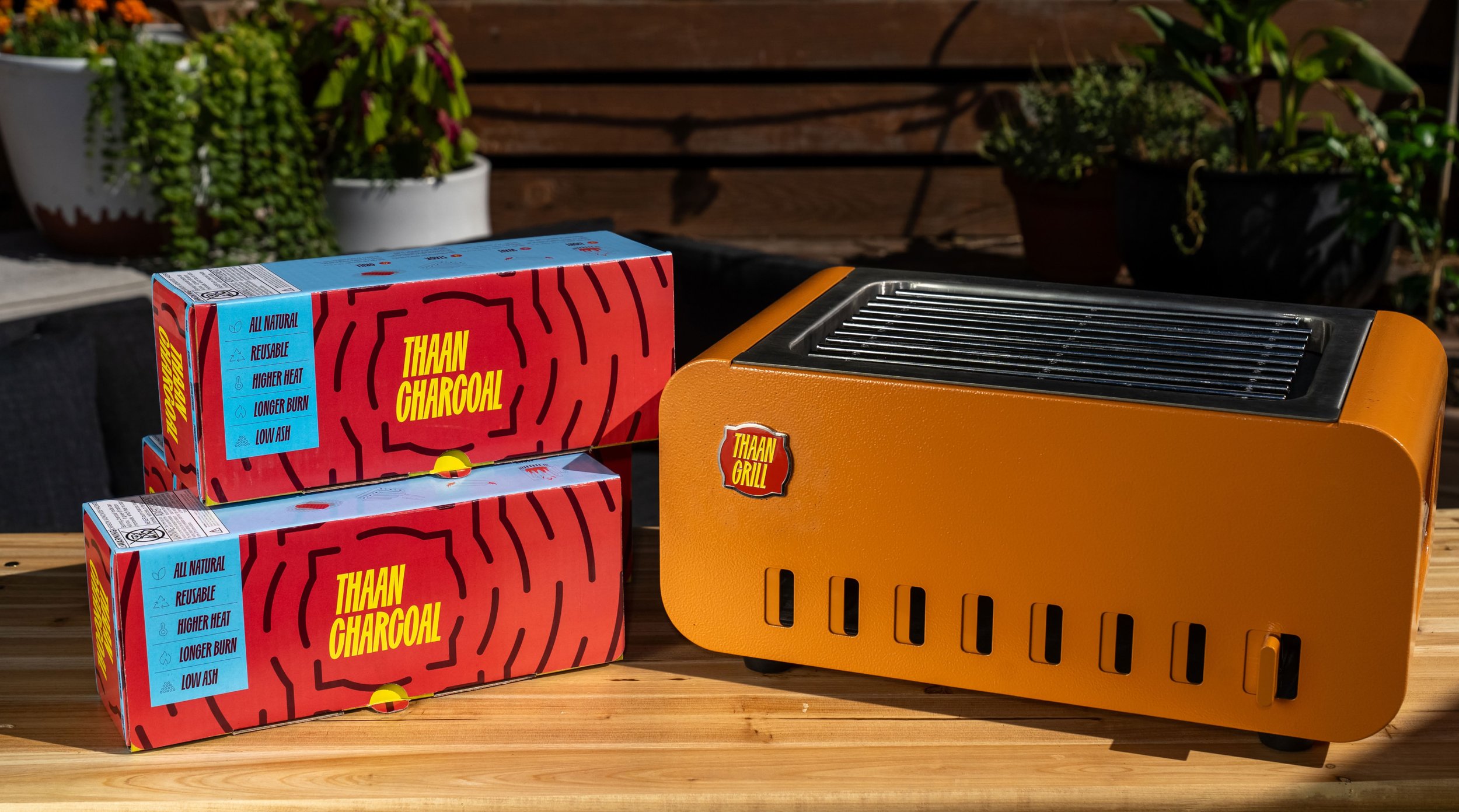 Boxes of charcoal labeled 'Thaan Charcoal,' plus a small orange grill with a black grate on top, set on a wooden surface outdoors with potted plants in the background.