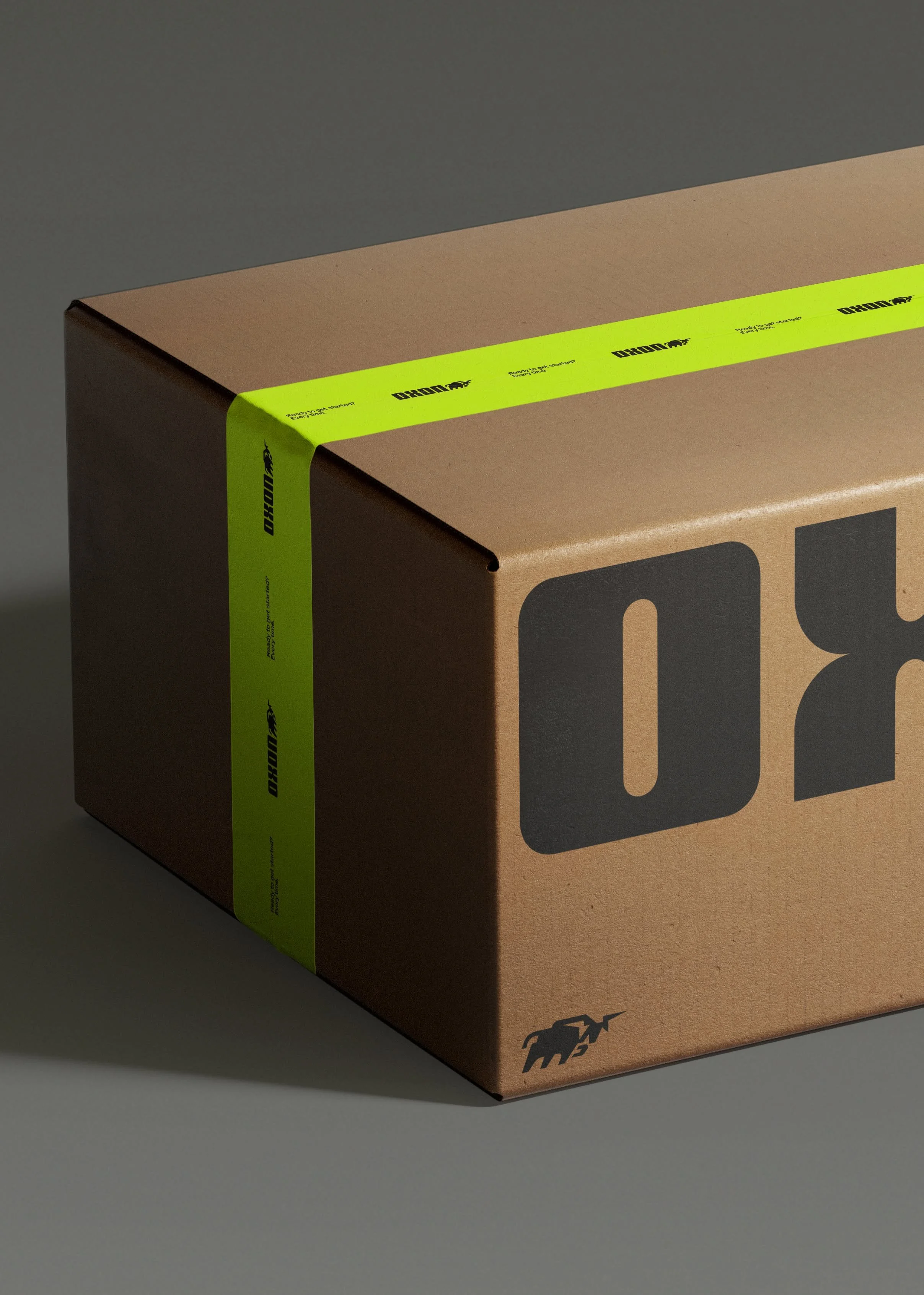 A cardboard box with a black and gray logo on the side, secured with bright yellow-green tape that has black text and logo. The box is placed on a gray surface with part of the shadow visible.