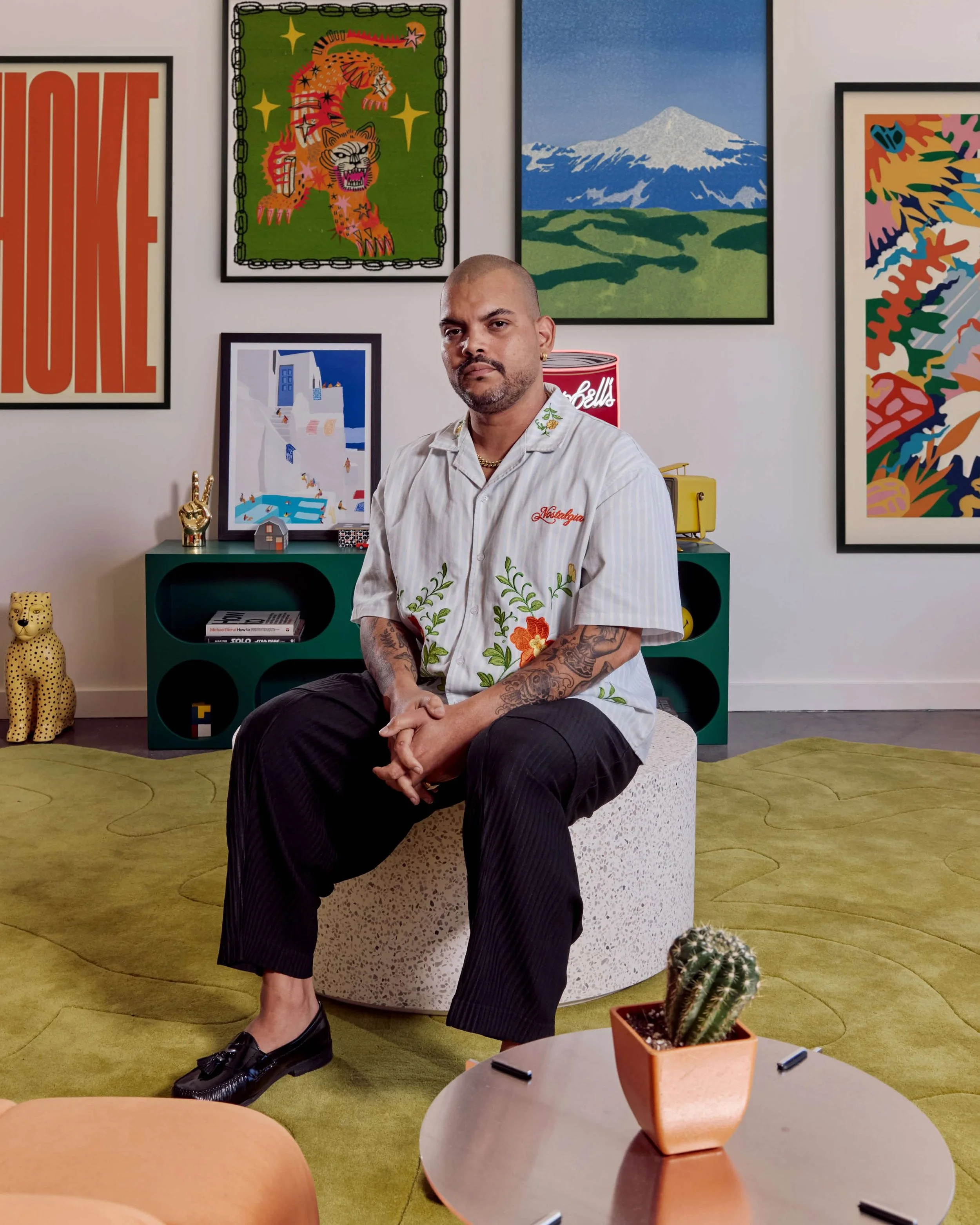 A man with a shaved head, tattoos, and a beard, wearing a light-colored shirt with green floral embroidery, seated on a white textured round stool in a modern, colorful room. Behind him are vibrant framed artworks, including a painting of a mountain, an abstract colorful piece, a tiger with stars, and a stylized landscape. There are decorative items like a gold hand sculpture, a Campbell's soup can lamp, a small house model, and a cheetah figurine on a green console. In the foreground, there is a white round table with a terracotta pot containing a cactus and several black markers. The room features a yellow carpet with a swirling pattern.