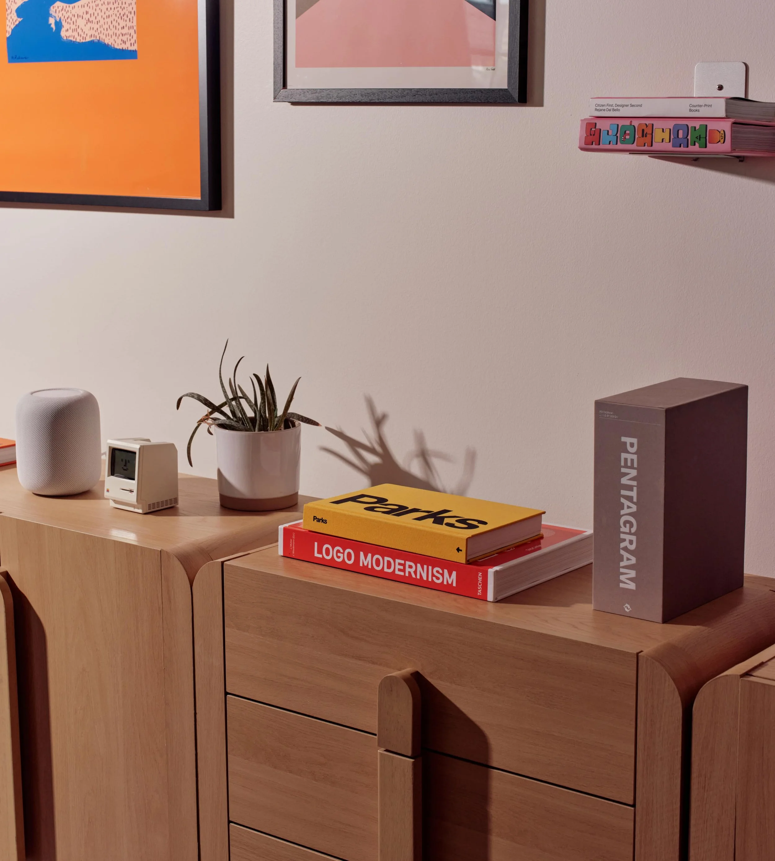 Wooden desk with a white smart speaker, a small digital clock, and a potted plant, with stacked books titled 'Parks,' 'Logo Modernism,' and 'Pentagram' on top and artwork on the wall behind.