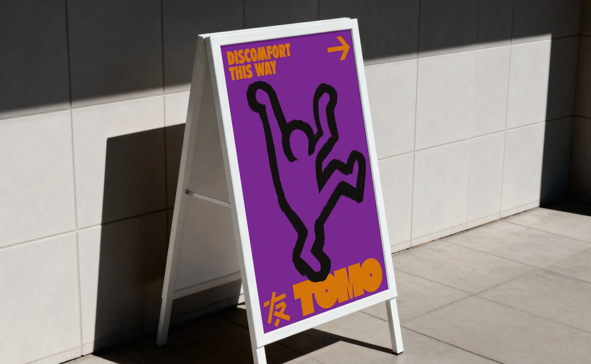 An outdoor A-frame sign with a purple background and black hand-drawn peace sign. The sign includes orange text reading "Discomfort This Way" and an orange arrow pointing to the right. It is positioned on a tiled sidewalk against a tiled wall.