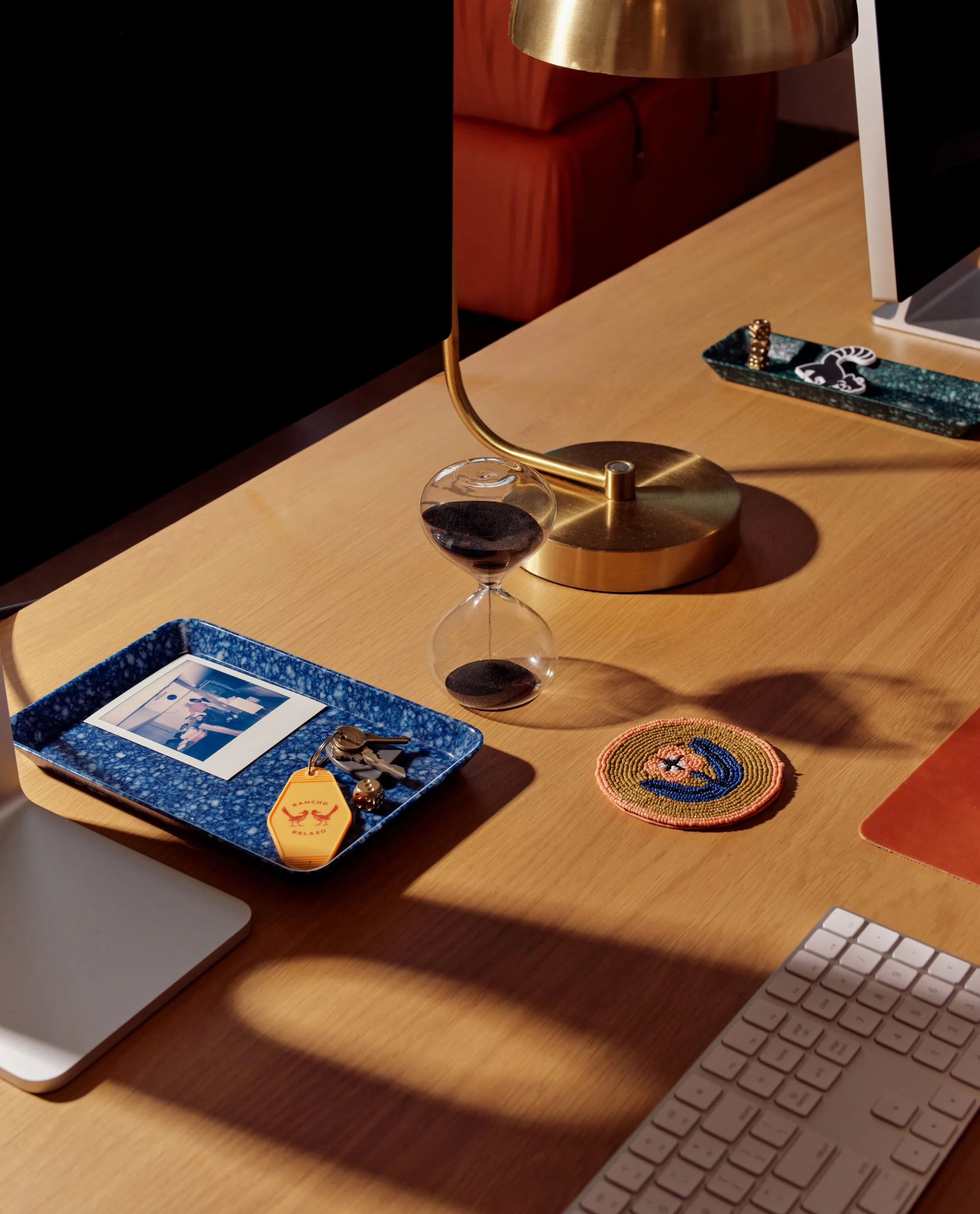 A wooden desk with a computer, a keyboard, a blue tray with a photo, a pair of keys, and decorative items including a beaded ring and a small woven coaster. Behind the desk, there is a gold lamp and a framed picture.