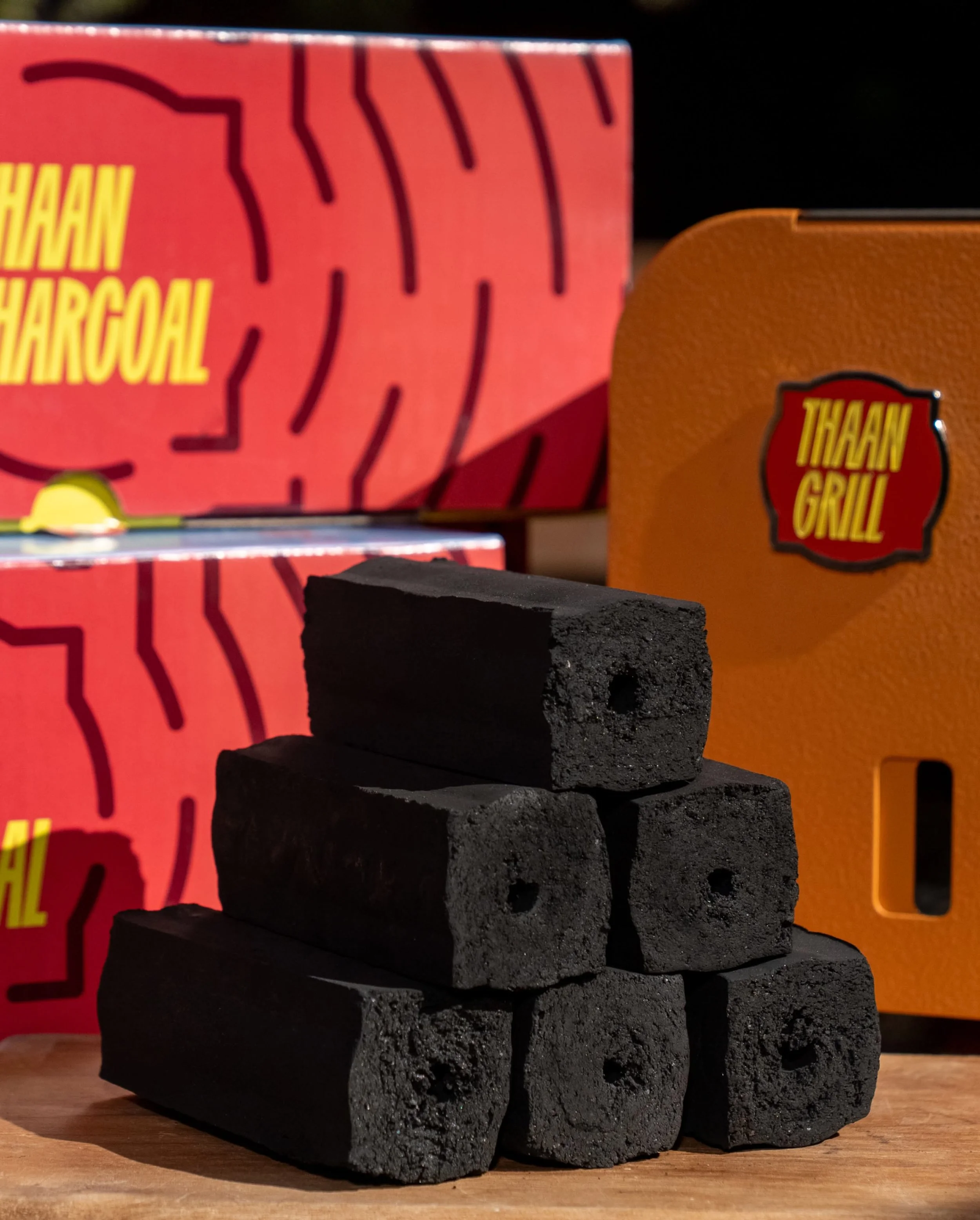 Stacked black charcoal briquettes with cardboard boxes labeled "Thaam Charcoal" and "Thaam Grill" in the background.