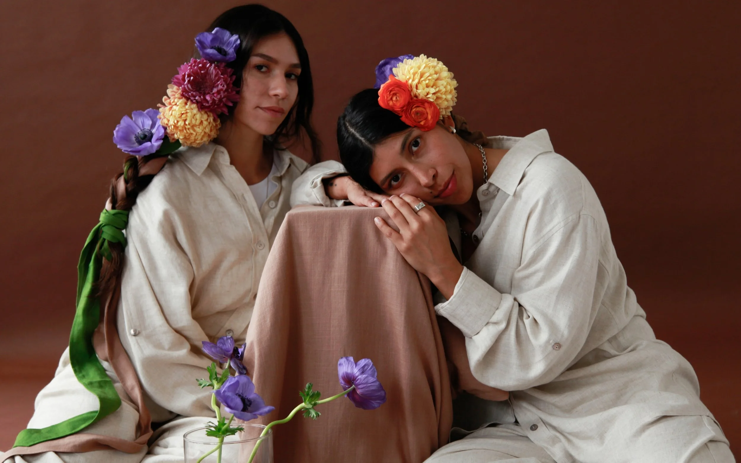 Two women with dark hair and light-colored shirts are posing together. They are decorated with colorful flowers on their heads and shoulders. One woman leans her head on a chair with a pinkish cover, and the other stands behind her. There is a glass vase with purple flowers in front of them.