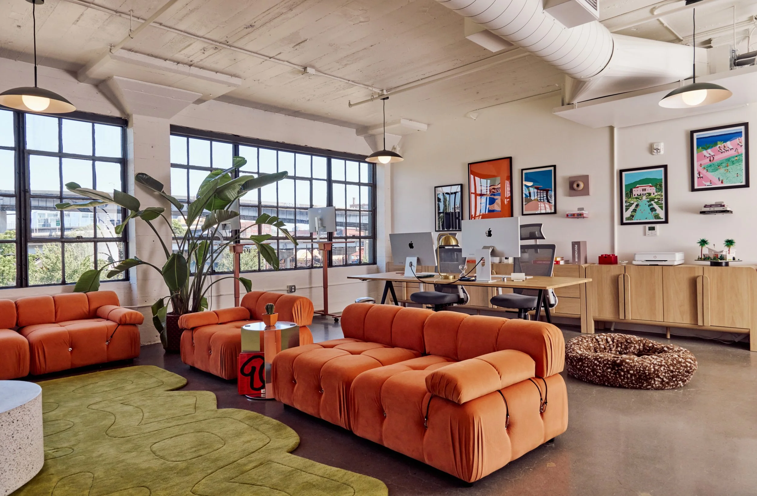 Modern office space with large windows, orange sofas, green area rug, plants, wall art, and workstations with computers.