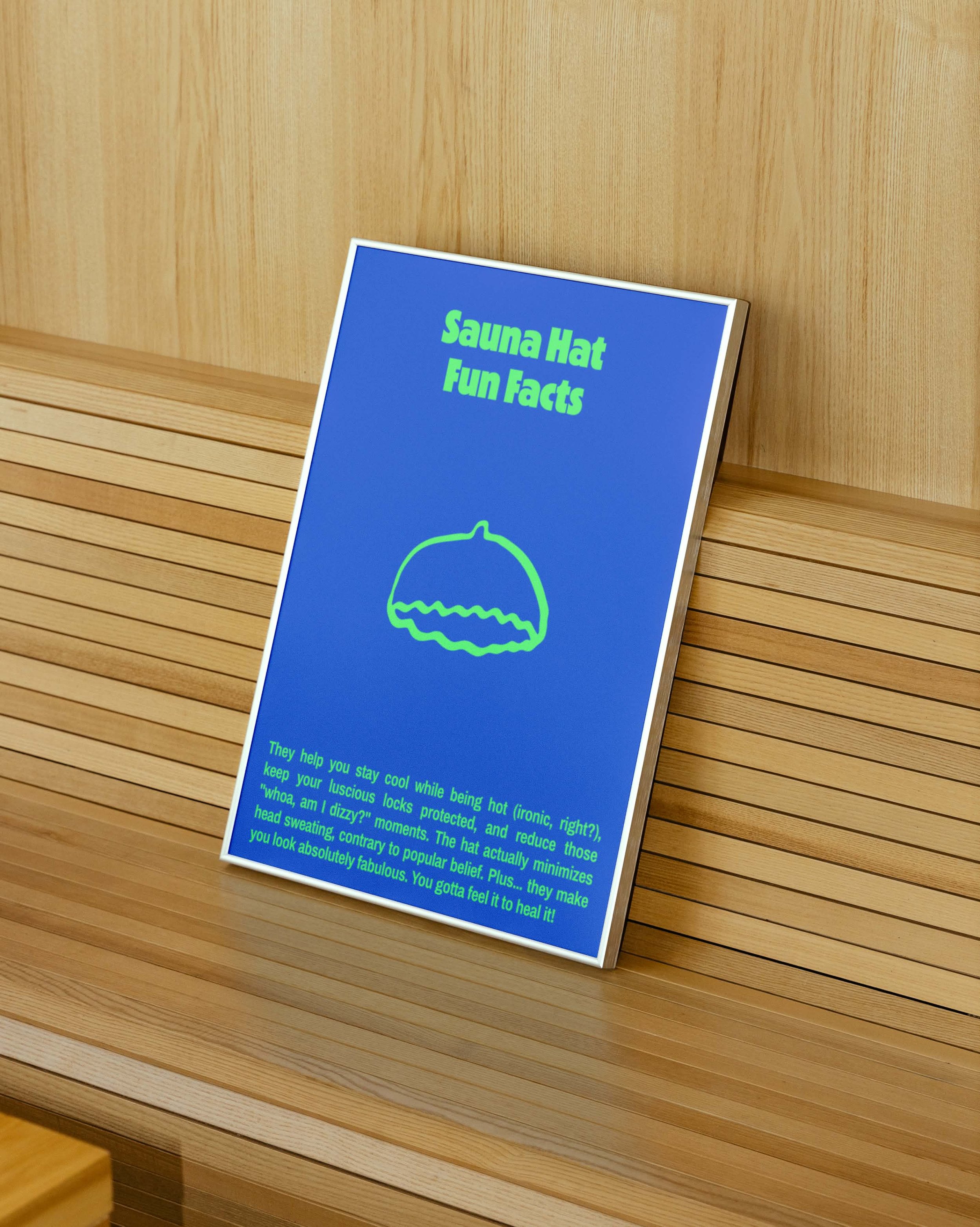 Signboard titled 'Sauna Hat Fun Facts' with a simple outline of a sauna hat and some text below explaining its benefits, placed on a slatted wooden surface.