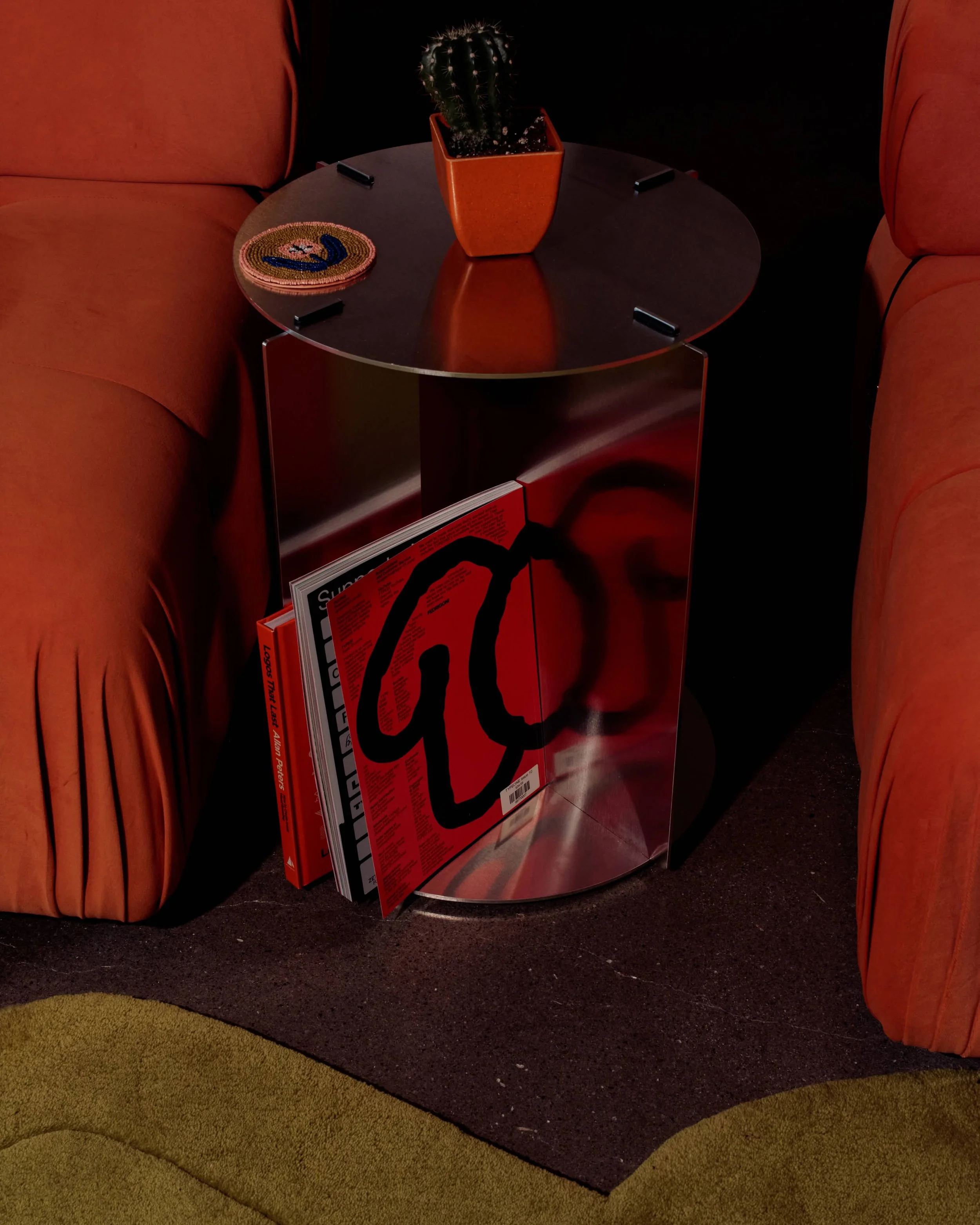 A round black table with a potted cactus in an orange square pot and a coiled magazine or newspaper with bold red, black, and blue colors on its cover. The table is between two orange upholstered chairs and has black clips attached to its edges.