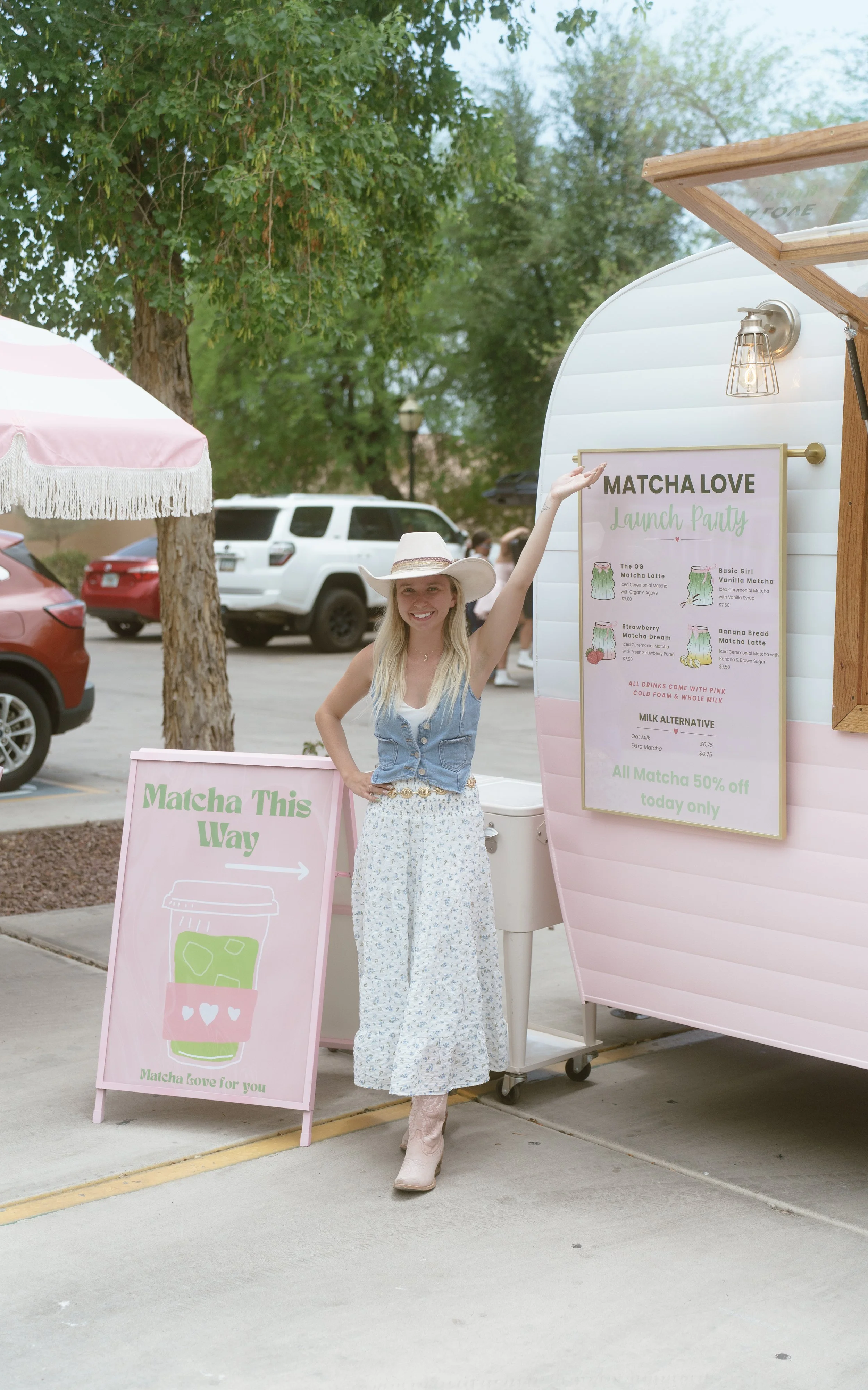 A woman wearing a cowboy hat, sleeveless denim vest, floral maxi skirt, and boots standing next to a pink and white mobile matcha tea stand, smiling and pointing at a menu sign.