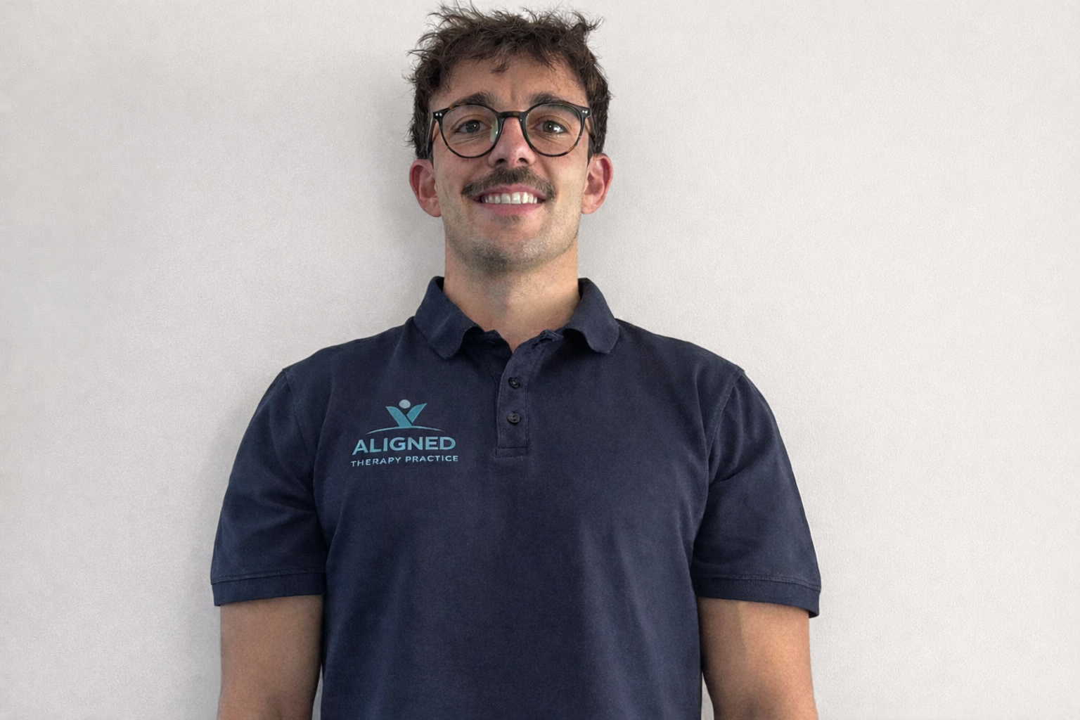 A man with curly brown hair, glasses, and a mustache, wearing a navy blue polo shirt with the logo 'Aligned Therapy Practice' standing against a plain white wall.