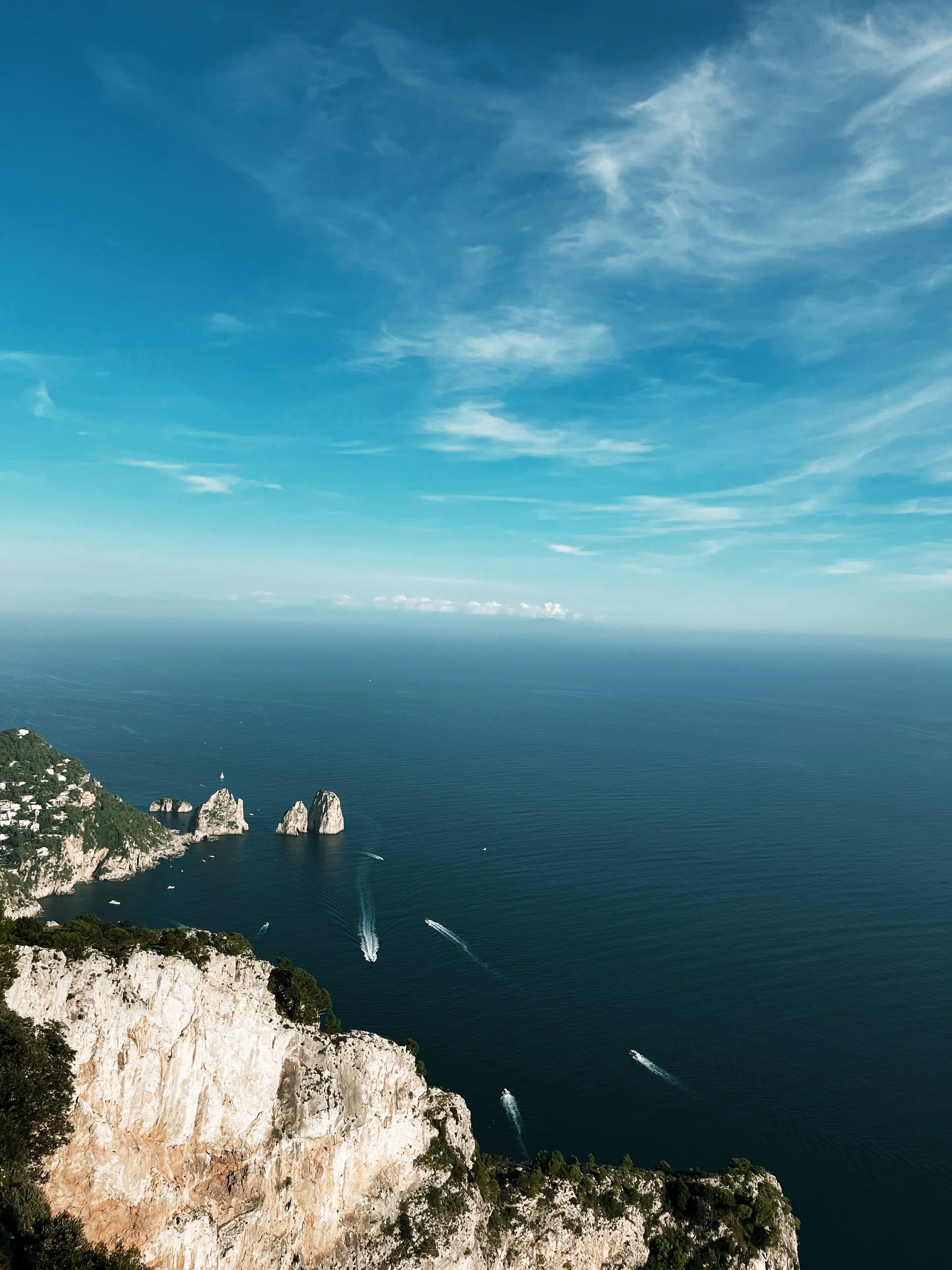 A coastal landscape featuring a rocky cliff with green vegetation in the foreground, steep white cliffs, a blue sea with boats creating white trails, and a bright cloudy sky.