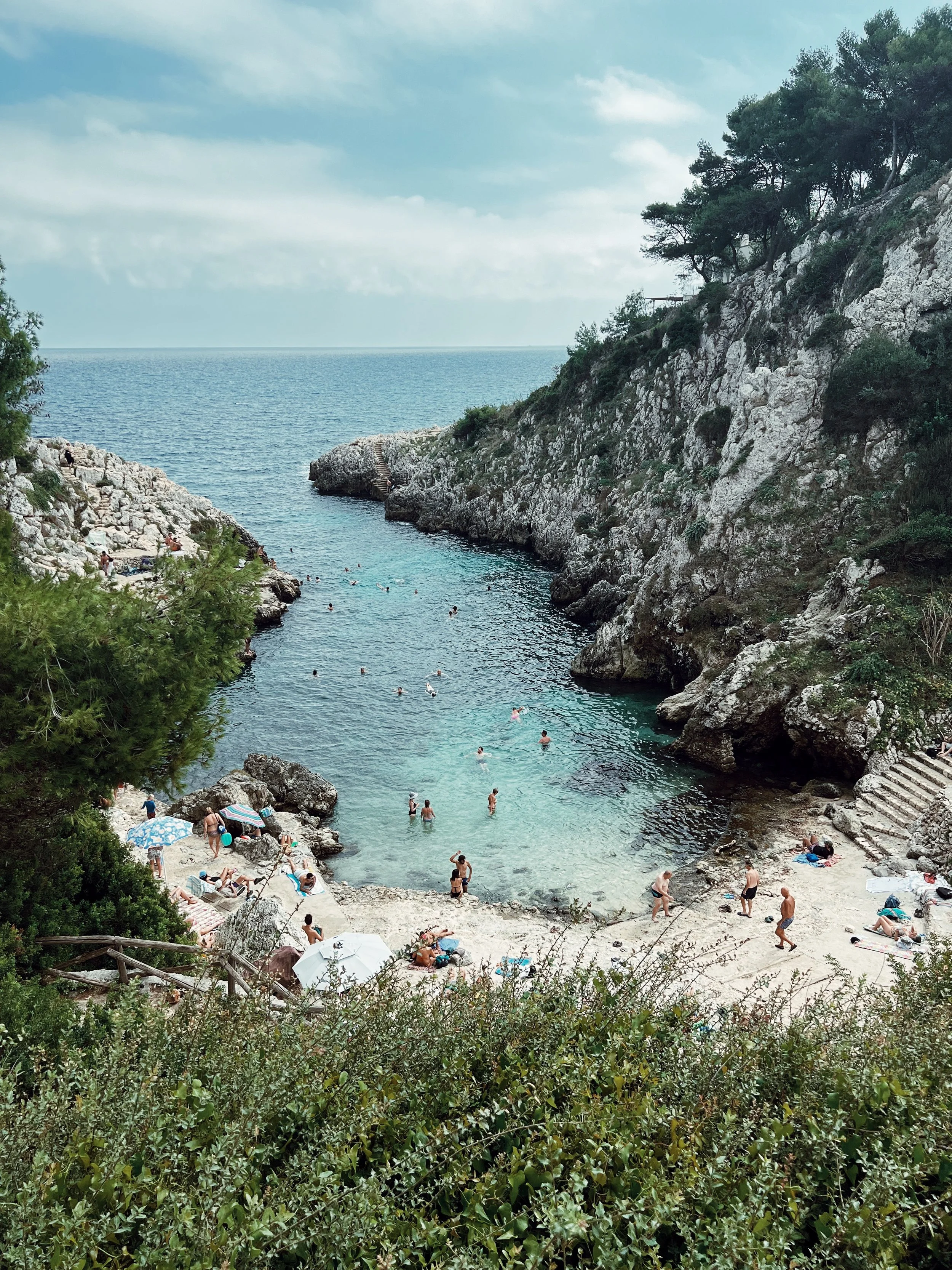 People swimming and relaxing at a small beach cove surrounded by rocky cliffs and green vegetation, with umbrellas and sunbathers on the sandy area.