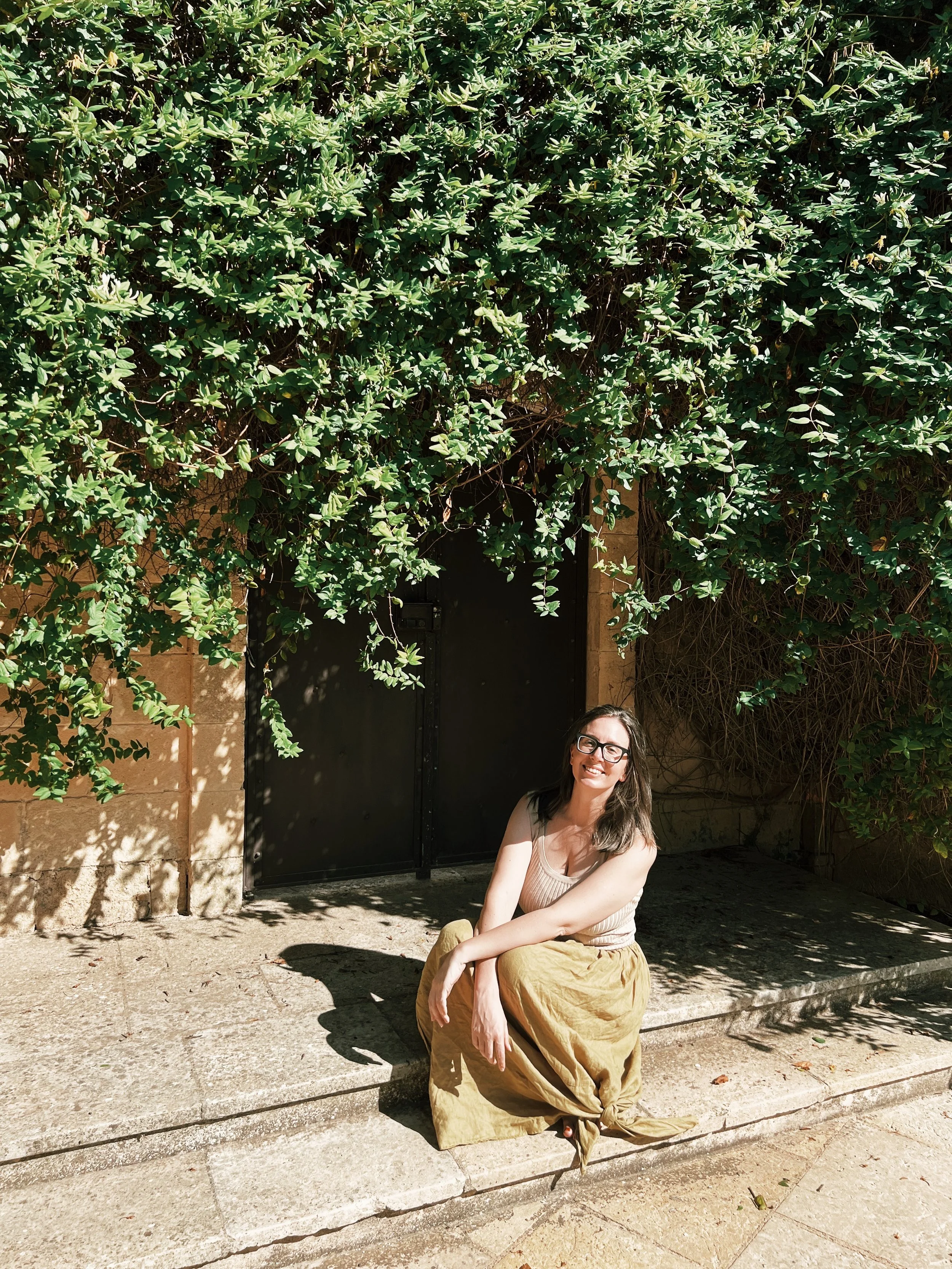 A smiling woman with glasses sitting on a curb in front of a large leafy green vine covering a wall and a black door.