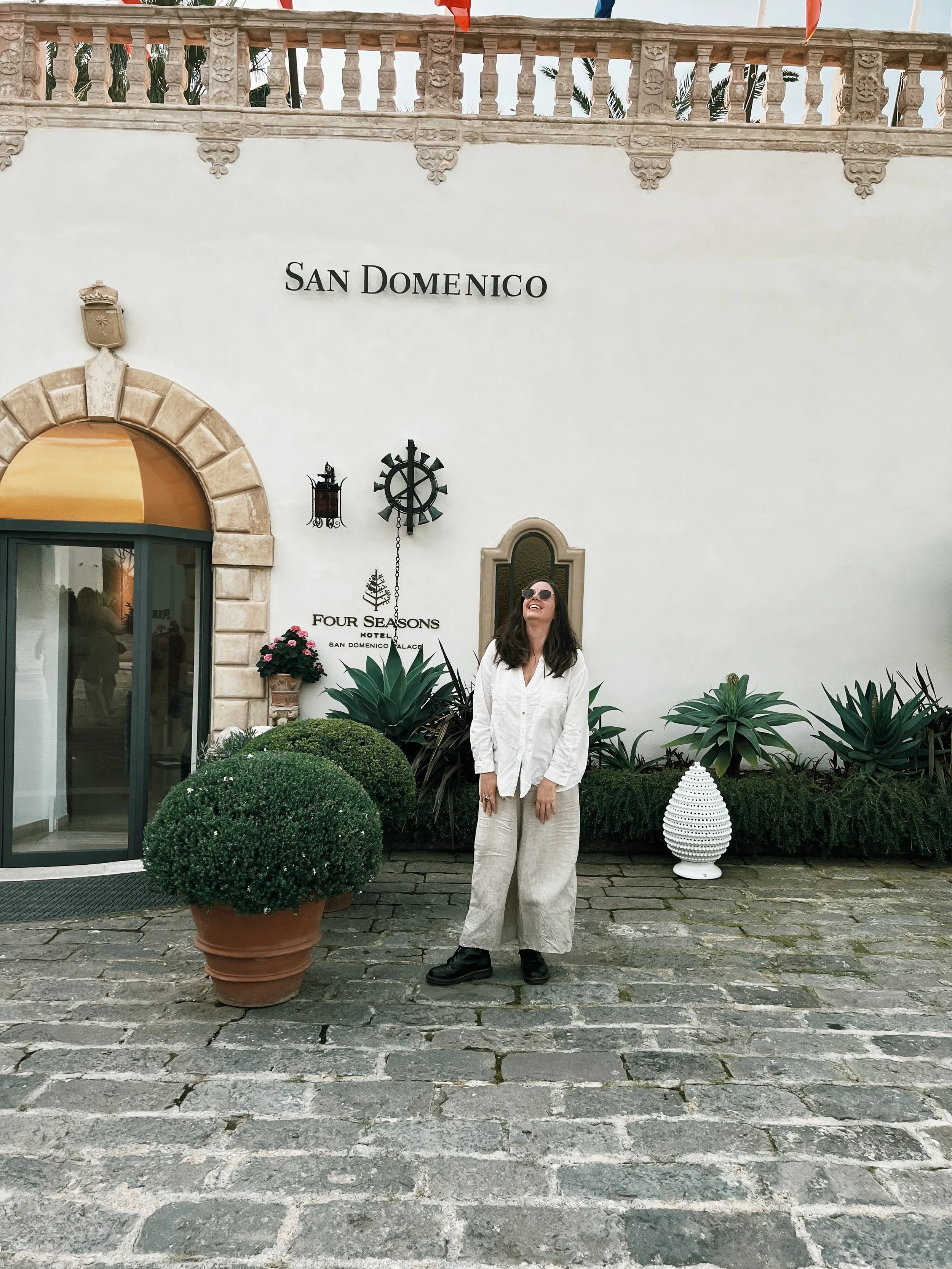 A woman with dark hair wearing sunglasses, a white shirt, and beige pants, stands outdoors in front of a white wall with the words 'San Domenico' and 'Four Seasons Hotel' on it. She is smiling and looking up, surrounded by various plants and decorative items, including a large potted plant, a white ceramic vase, and a dark metal wall clock.