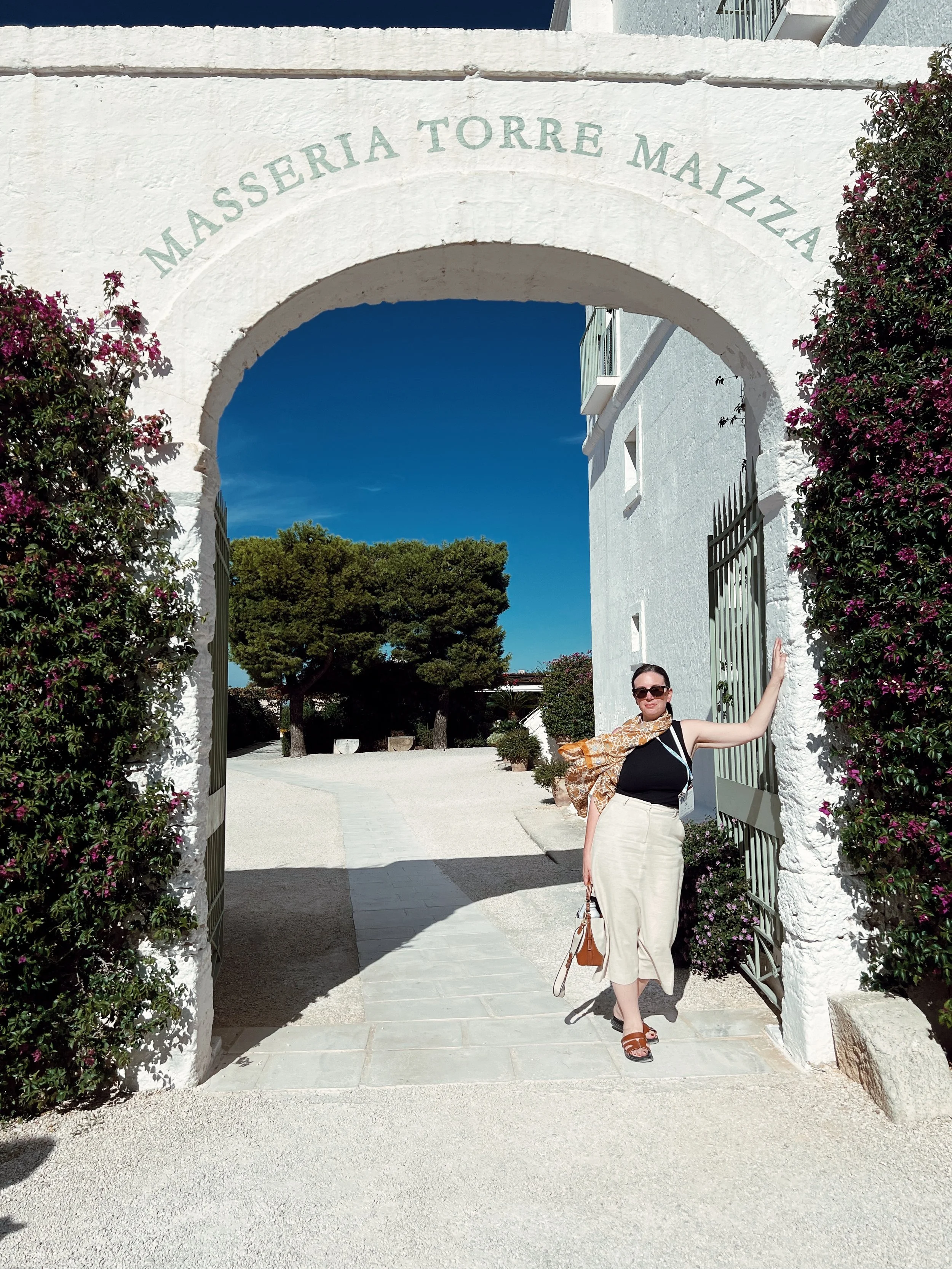 A woman standing under a white archway with the words "Masseria Torre Maizza" inscribed above. She is wearing sunglasses, a black top, beige skirt, brown sandals, and is holding a tan handbag. There are pink flowering bushes on either side of the arch and trees in the background under a clear blue sky.