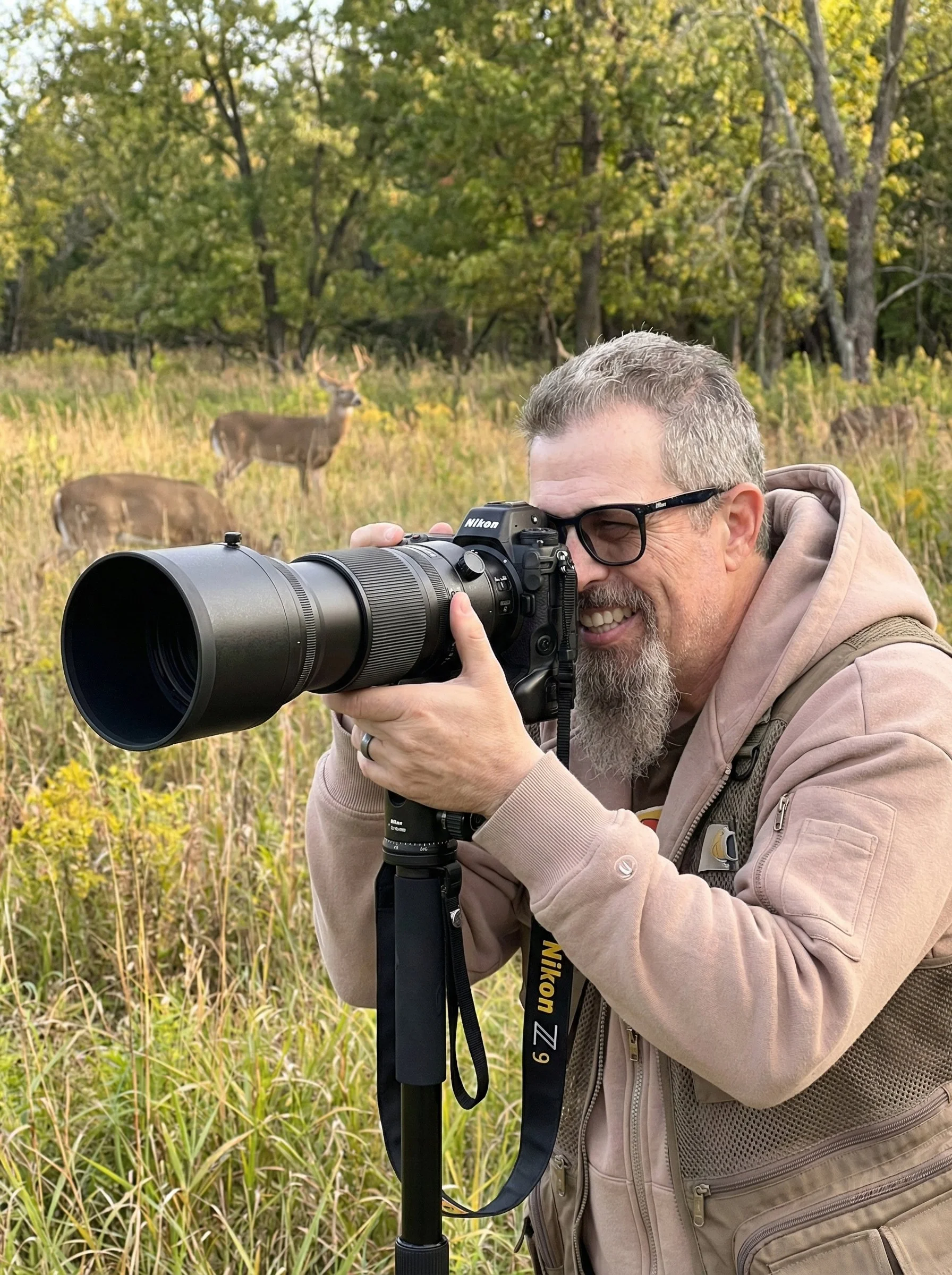 A man with glasses, a beard, and a beige jacket is taking a photograph with a Nikon camera mounted on a tripod in a grassy field. Deer are visible in the background among trees.