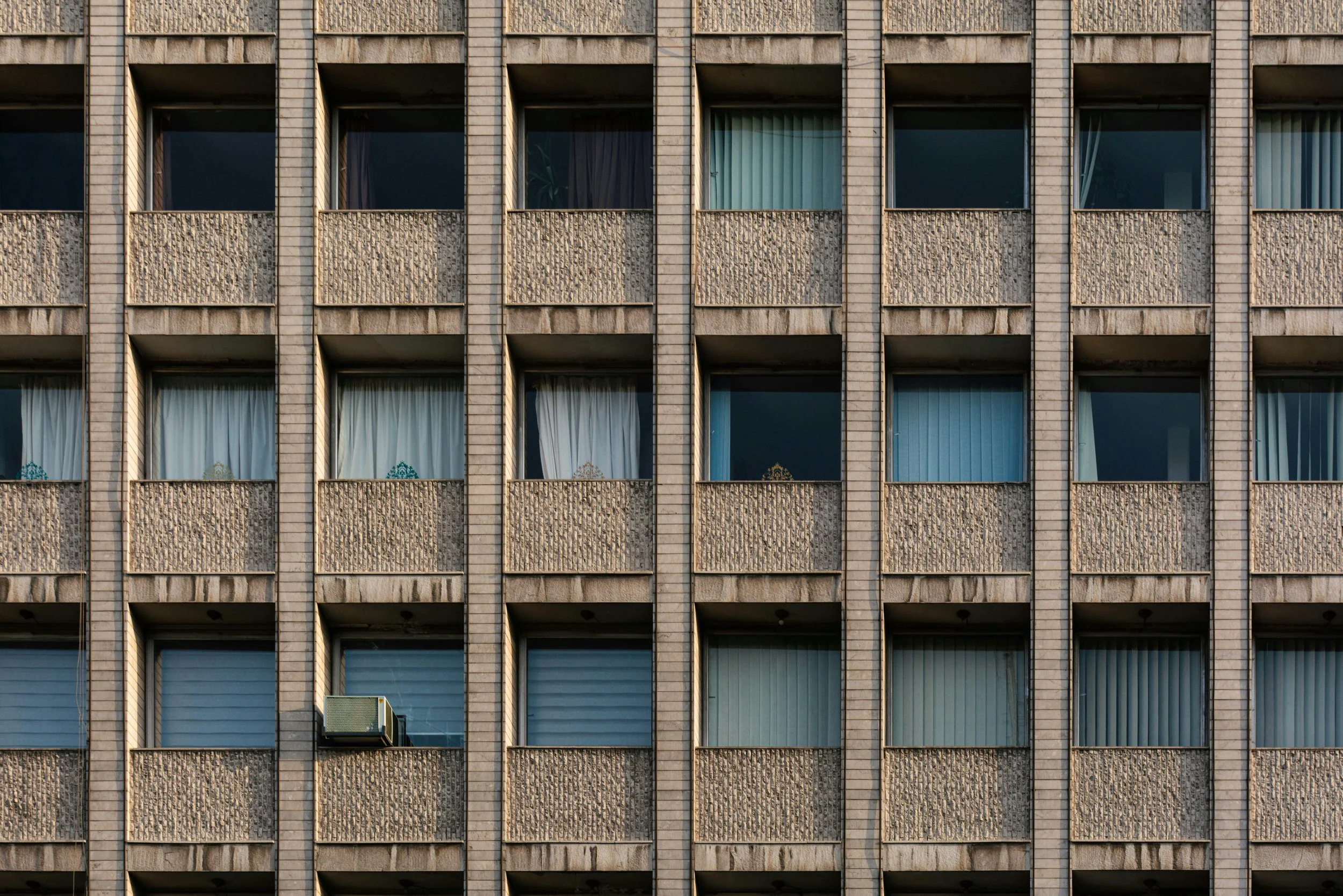 Close-up of a multi-story apartment building facade with numerous windows, some with curtains and one with an air conditioning unit.