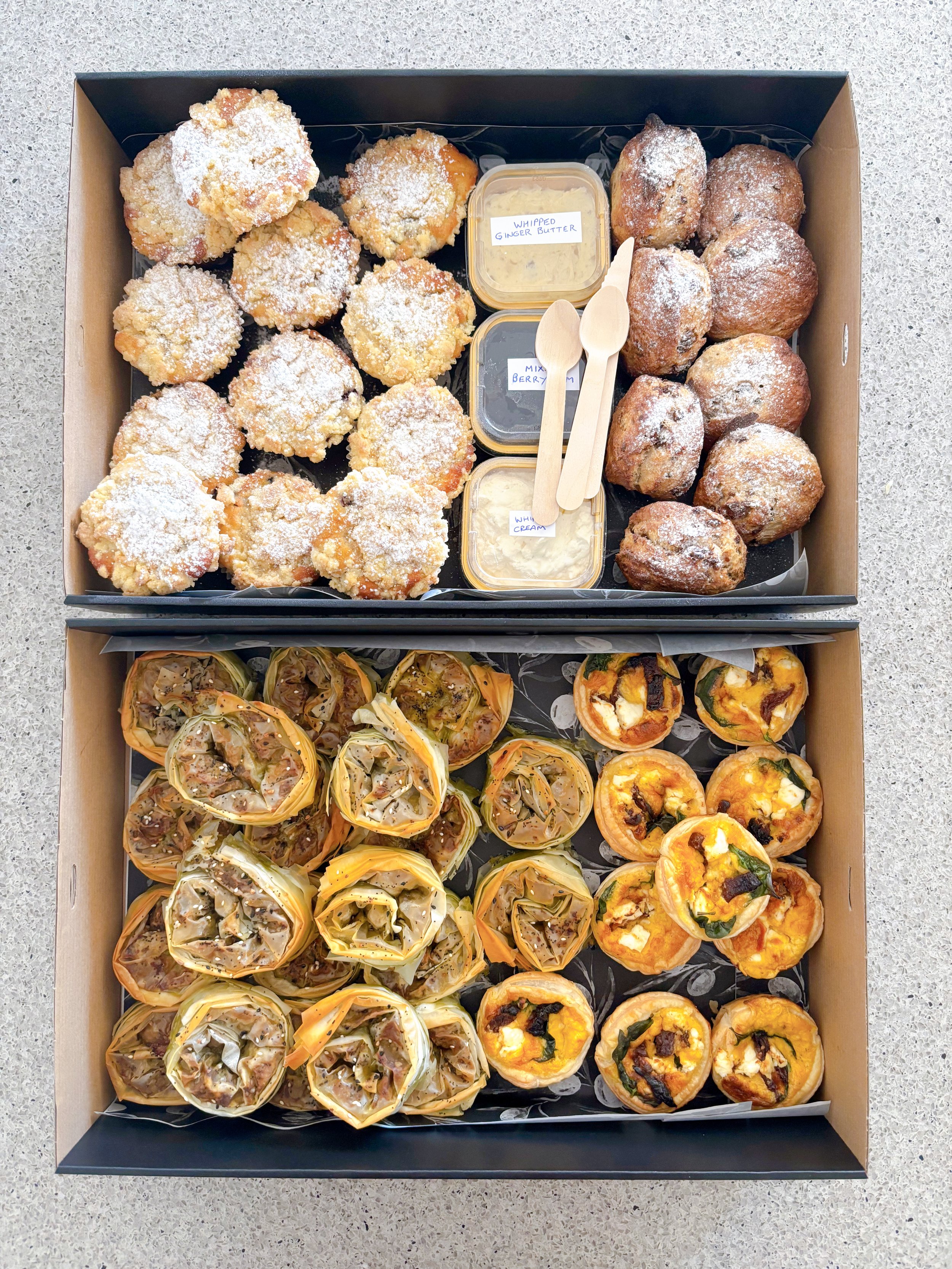 Two boxes of assorted baked goods: the top box scones muffins and small containers of whipped ginger butter, berry compote, and whipped cream with wooden spoons; the bottom box contains individual servings of filo pastries and mini savory pies.