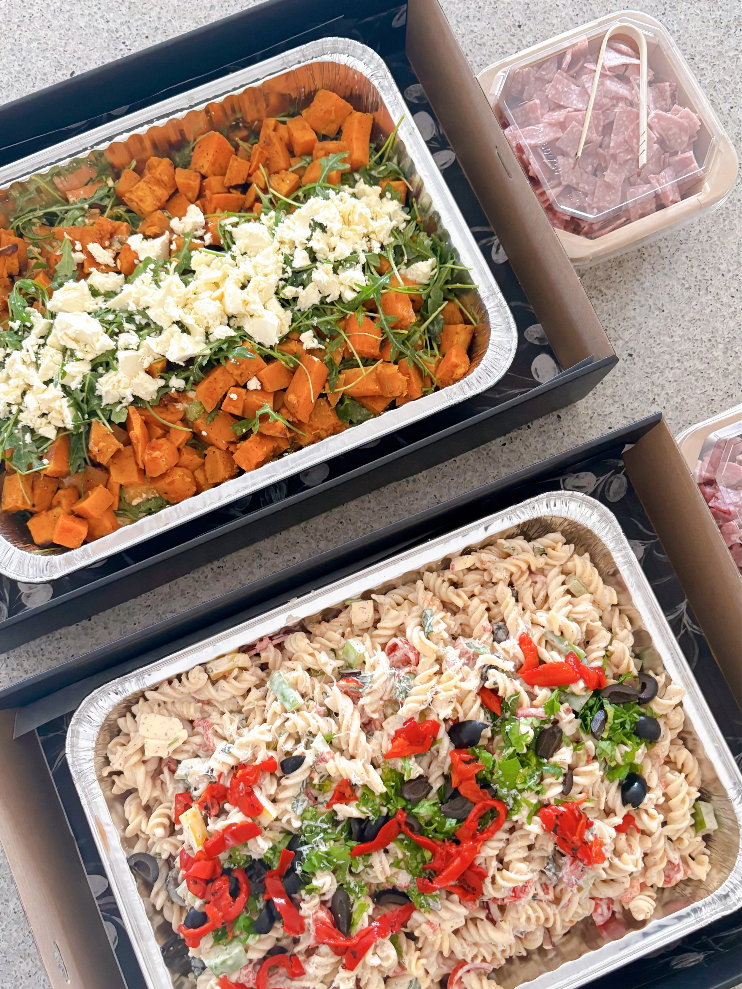 Two aluminum trays of pasta salads with various toppings, one with chopped sweet potatoes and feta cheese, and the other with rotini pasta, black olives, chopped red peppers, and shredded chicken.