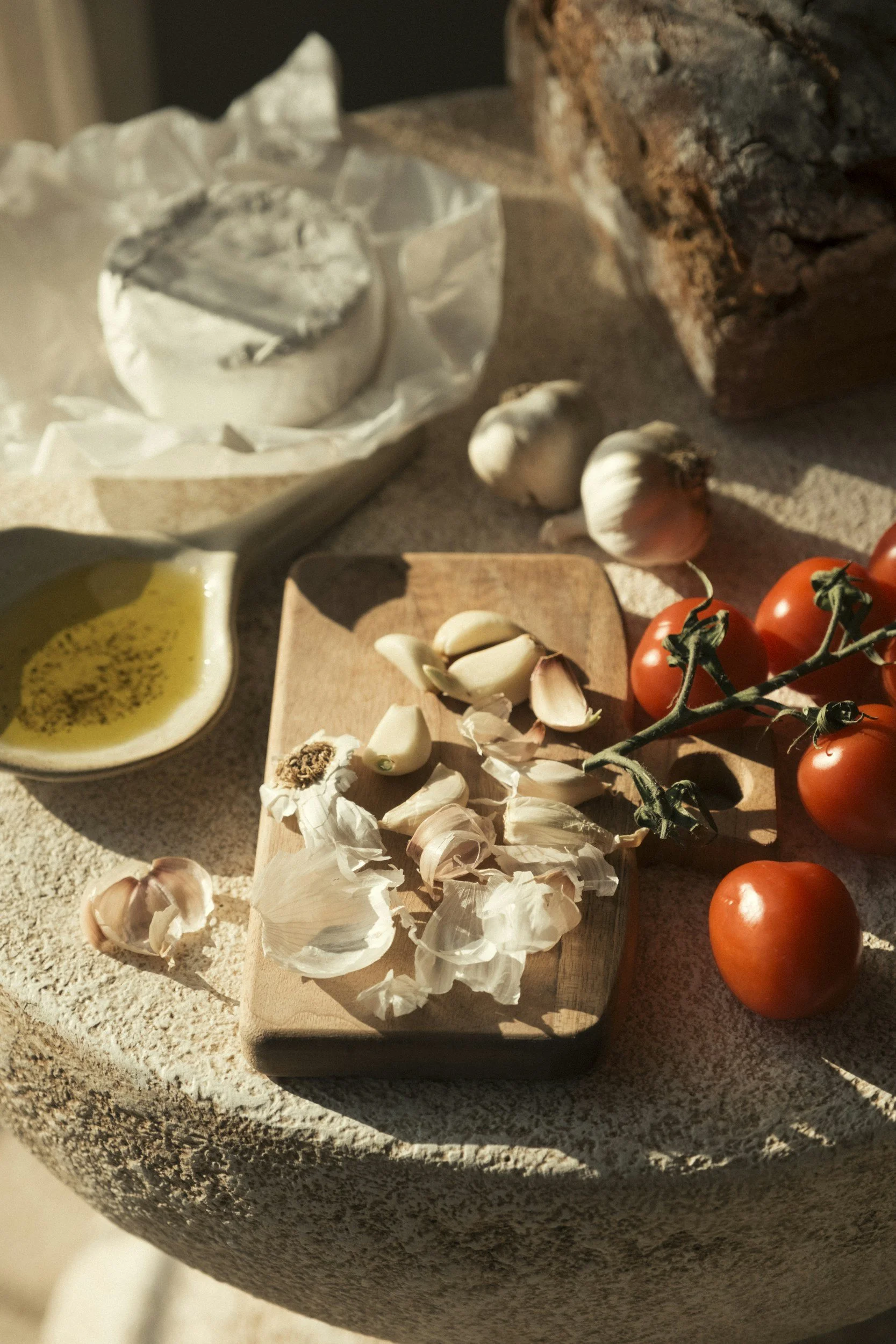 Fresh garlic, cherry tomatoes on the vine, and garlic cloves on a wooden cutting board, with garlic bulb, garlic powder, and olive oil nearby on a rustic surface.