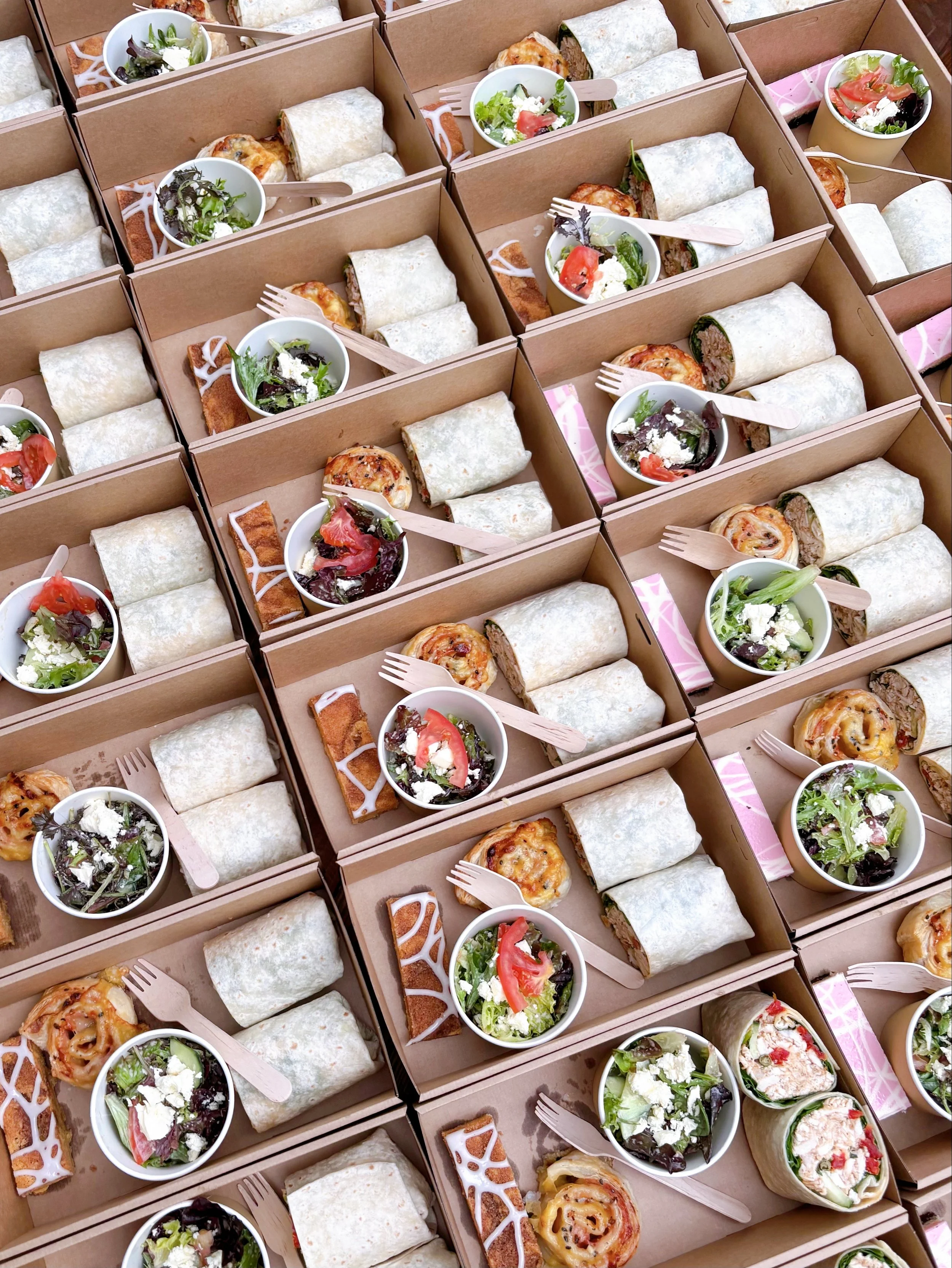 Multiple meal boxes containing wraps, small salads, and baked items, with some boxes showing cookies and utensils.