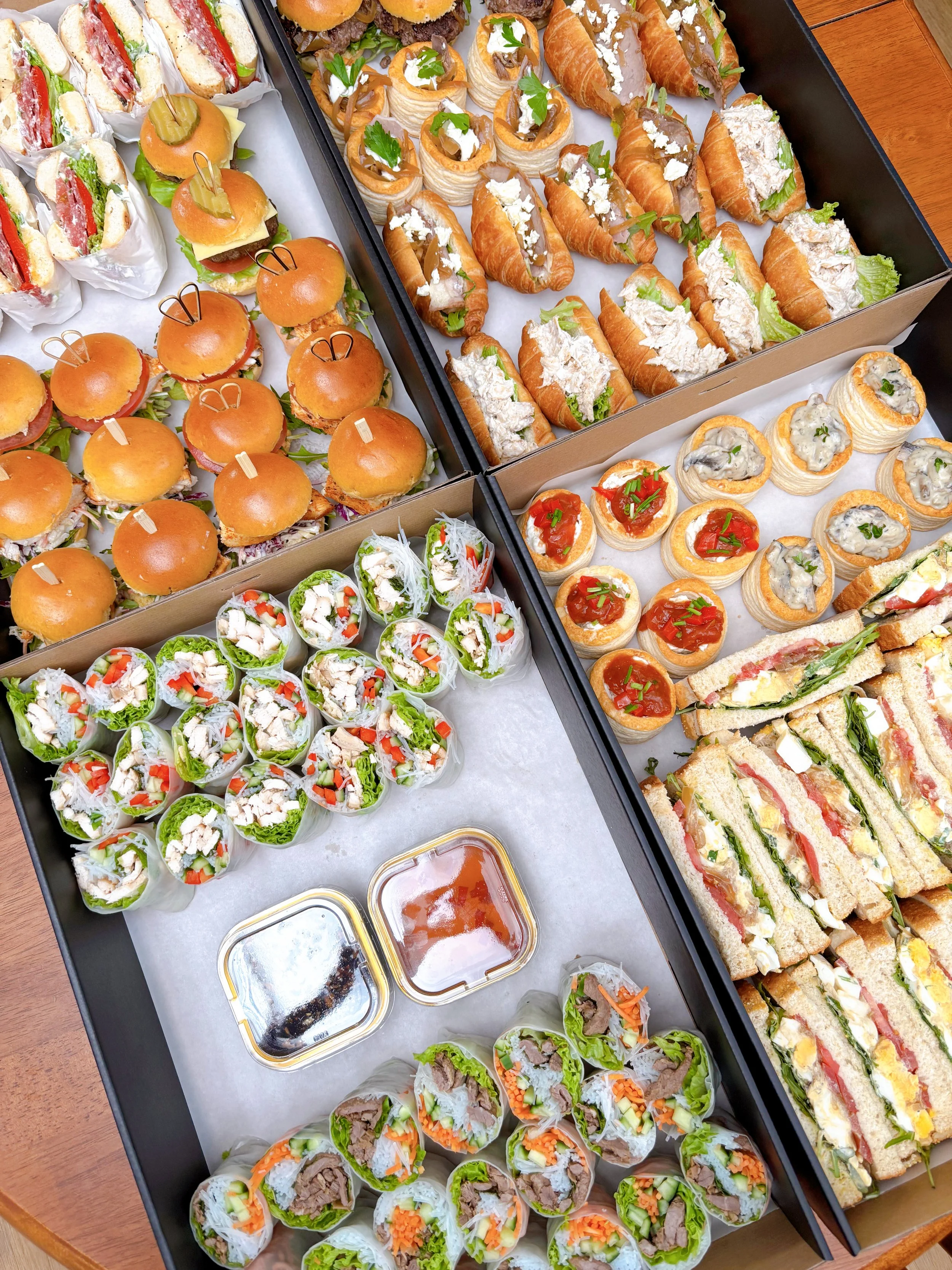 Tray of assorted finger foods including mini sandwiches, chicken wraps, spring rolls, croissants with fillings, and small appetizers, with dipping sauces at the bottom.