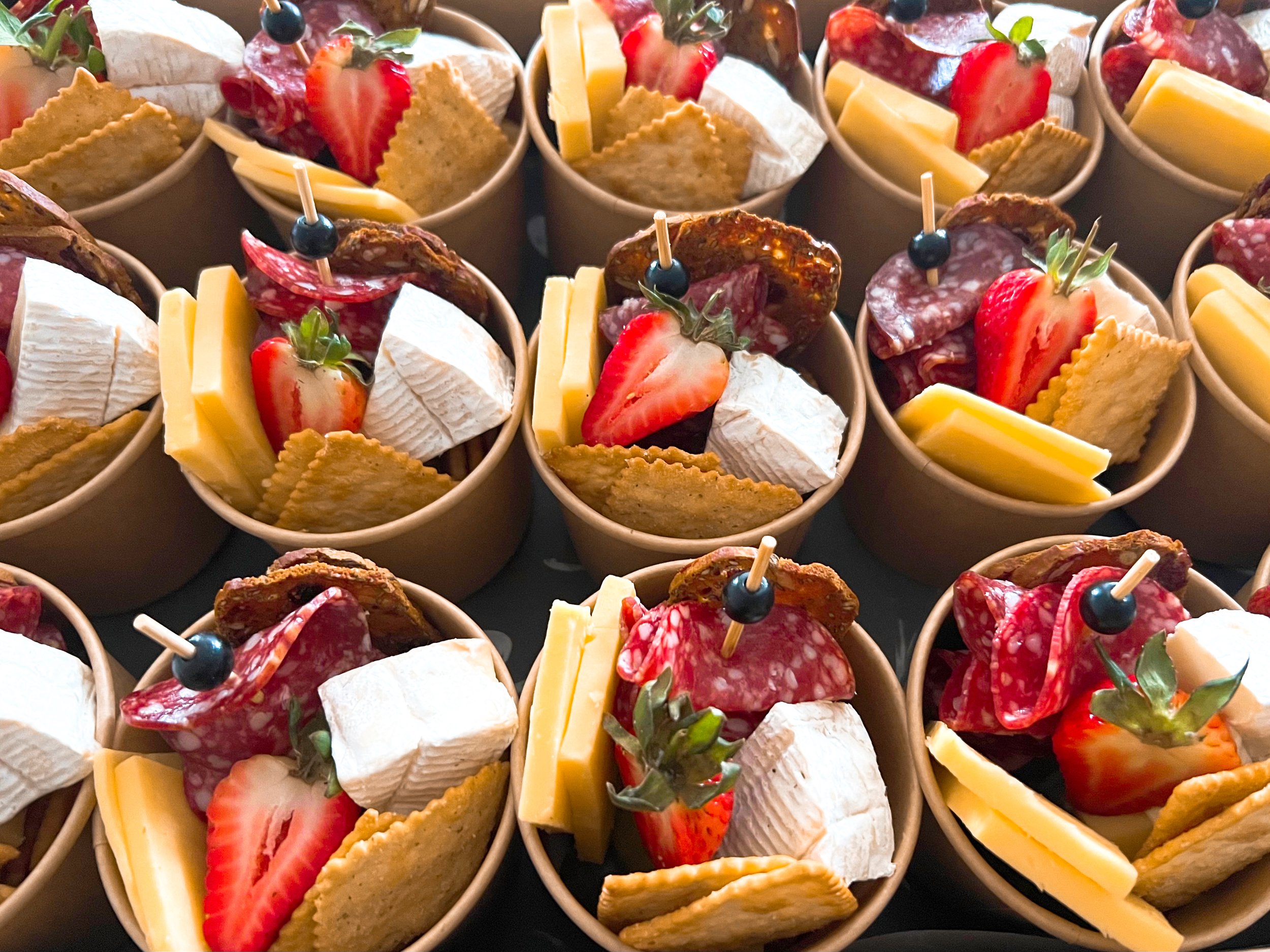 Small cups filled with assorted cheeses, strawberries, slices of cured meats, crackers, and garnished with fresh herbs and blueberries.